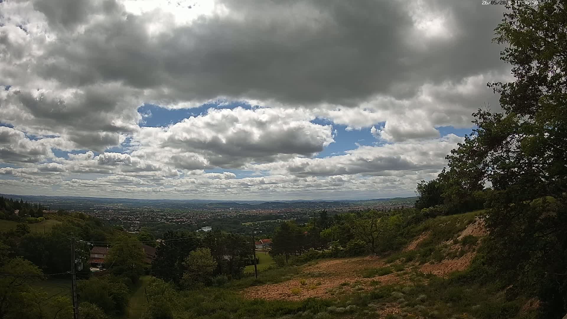 A partly cloudy sky overlooks a hilly landscape with green trees and a distant city visible under a mostly cloudy sky.