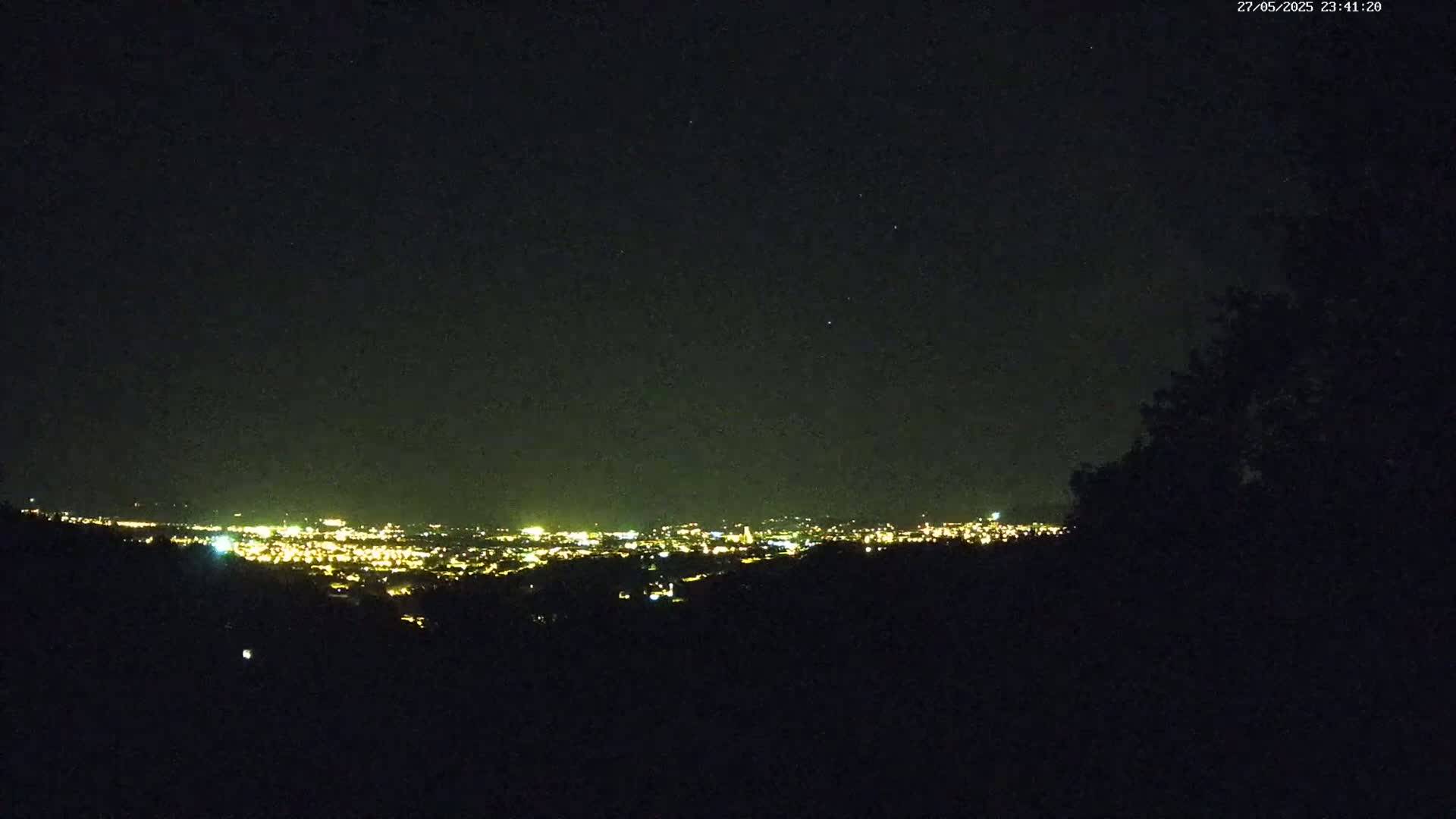 A nighttime view from a hilltop shows a distant city glowing with lights under a clear, starry sky.