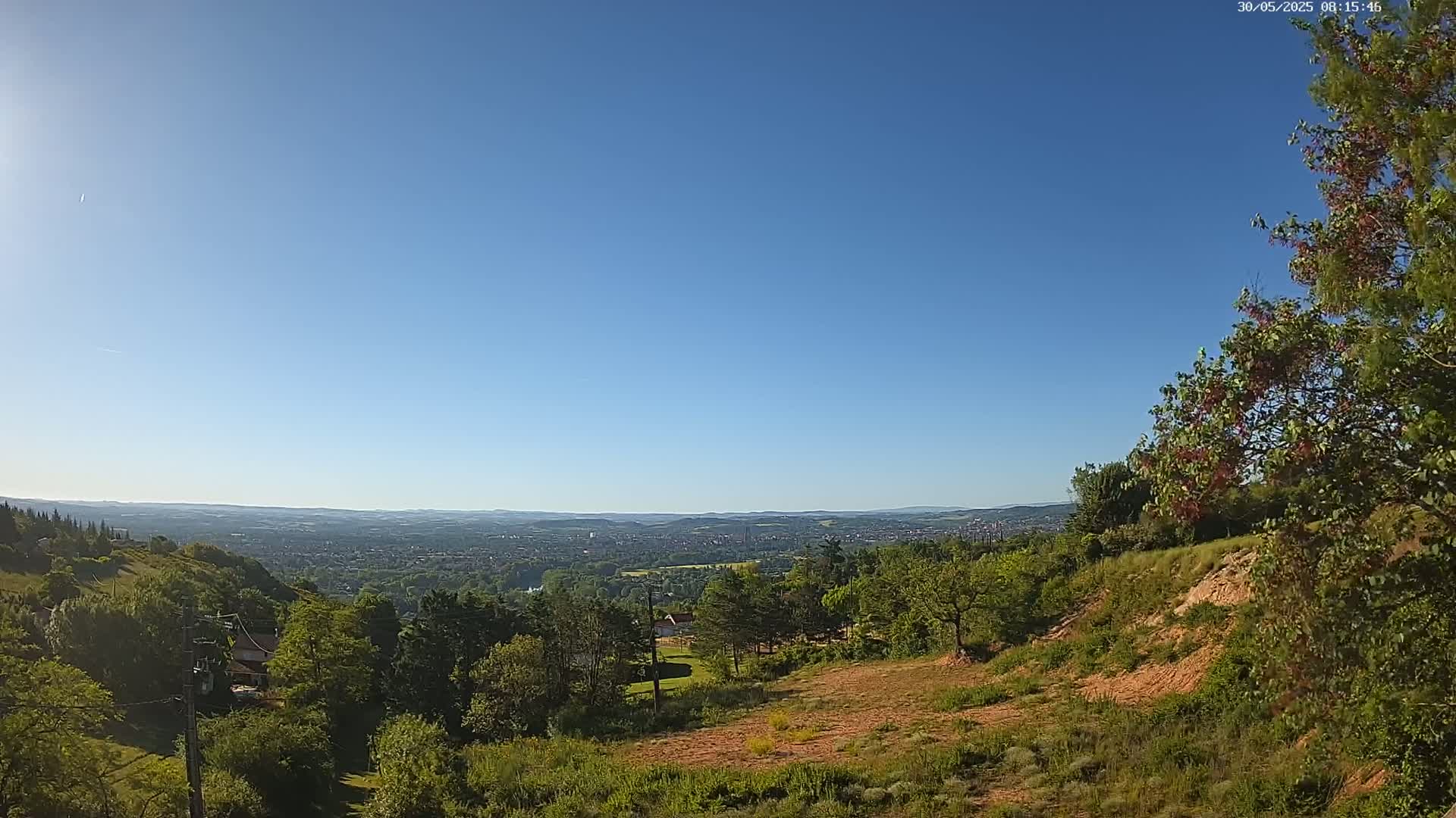 A high-angle, long shot of a hilly landscape on a clear, sunny day shows a distant city spread across a valley, surrounded by green trees and vegetation.