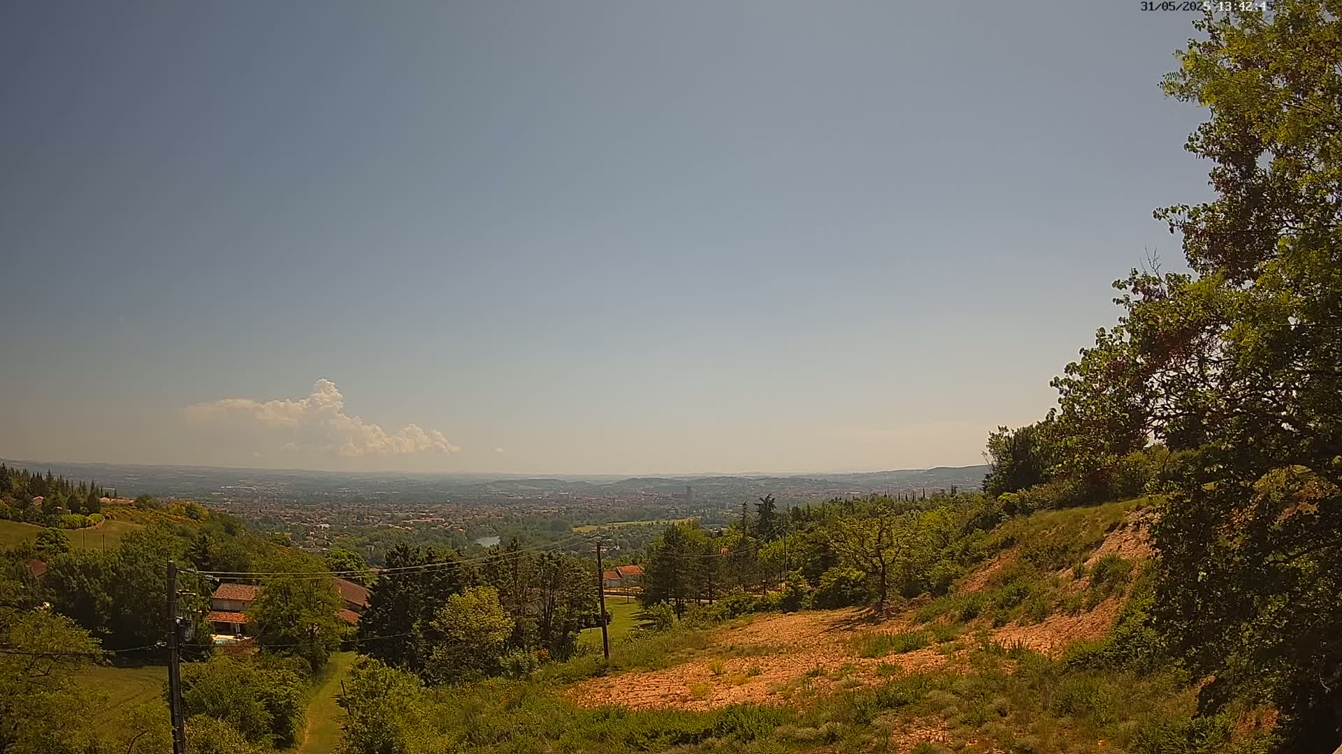 A hilly, wooded landscape under a mostly clear, sunny sky overlooks a distant city.