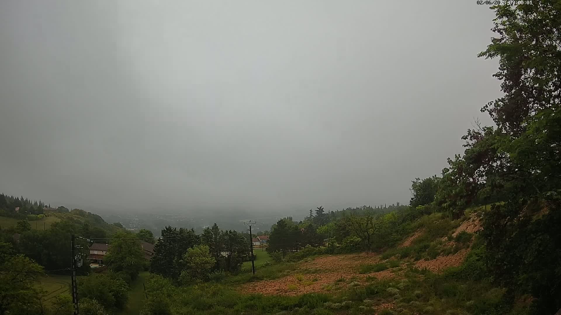 A foggy, overcast day reveals a hilly landscape with lush green vegetation, a few houses, and a distant view of a town or city in the valley below.