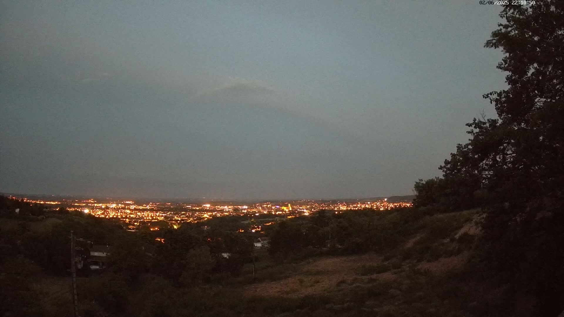 A nighttime view from a hilltop shows a city glowing with lights under a dusky, overcast sky.