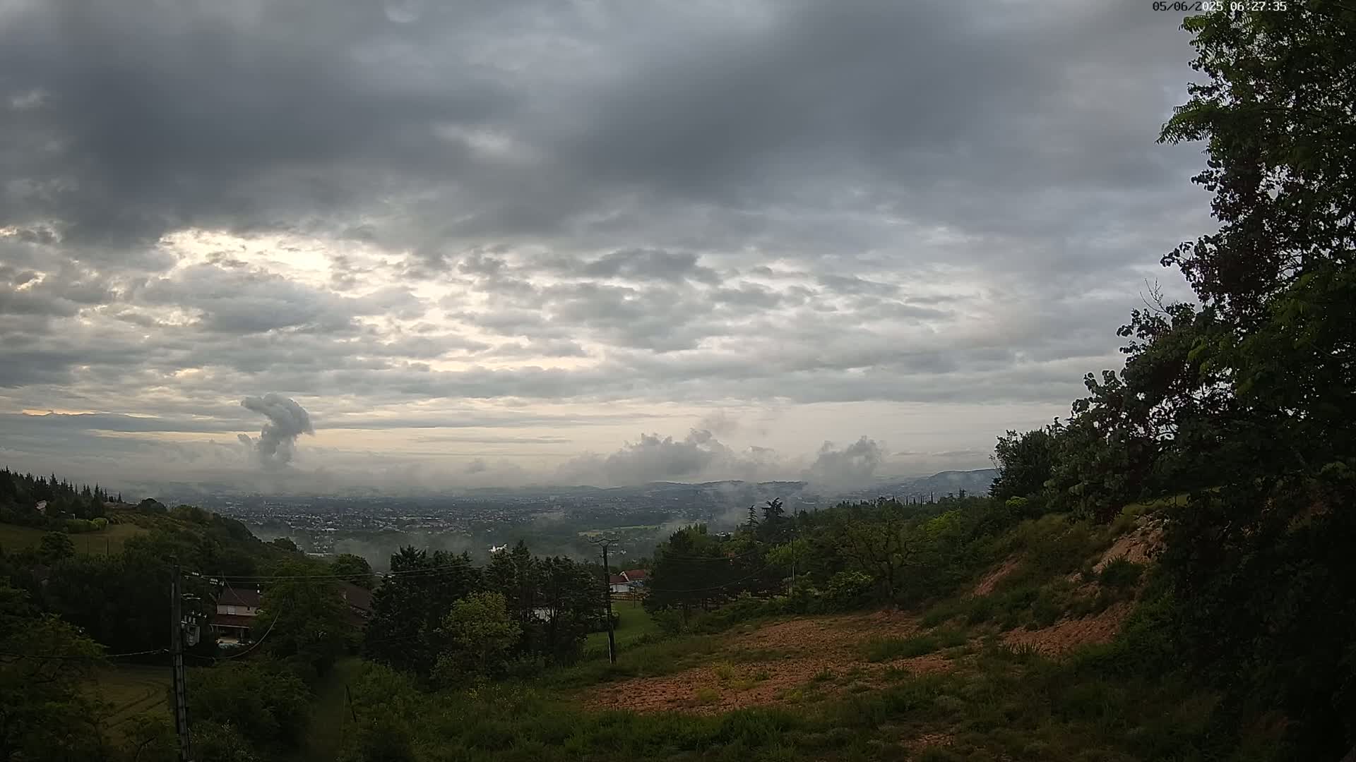 A view from a hillside shows a valley town covered in morning mist under a cloudy sky.