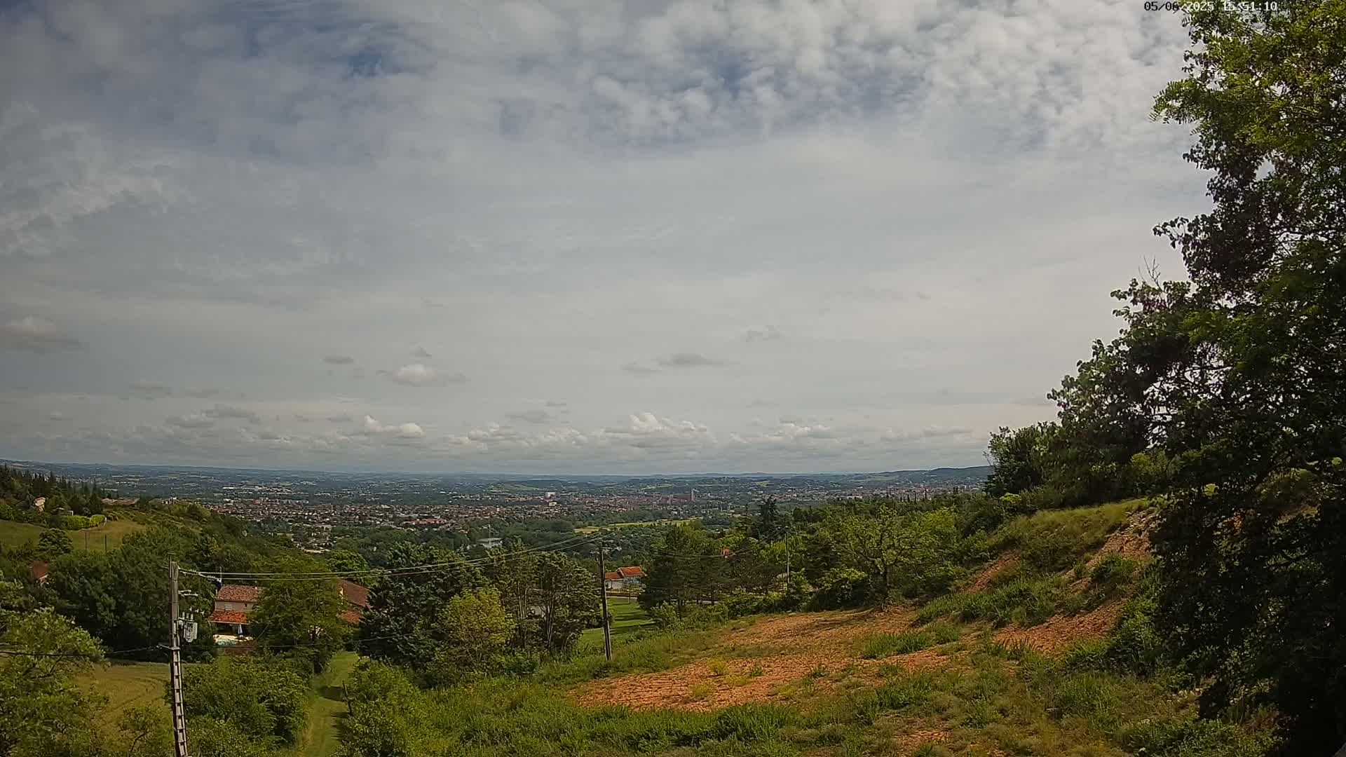 A partly cloudy day reveals a sprawling city view from a hilly, wooded vantage point with sparsely vegetated, reddish-brown earth visible in the foreground.
