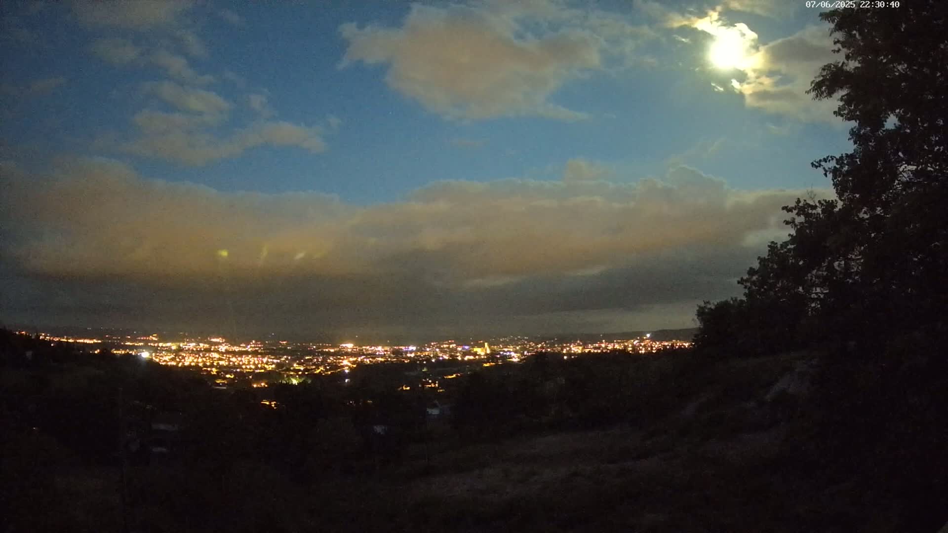 A nighttime view of a city illuminated below, partially obscured by a layer of clouds under a bright moon.
