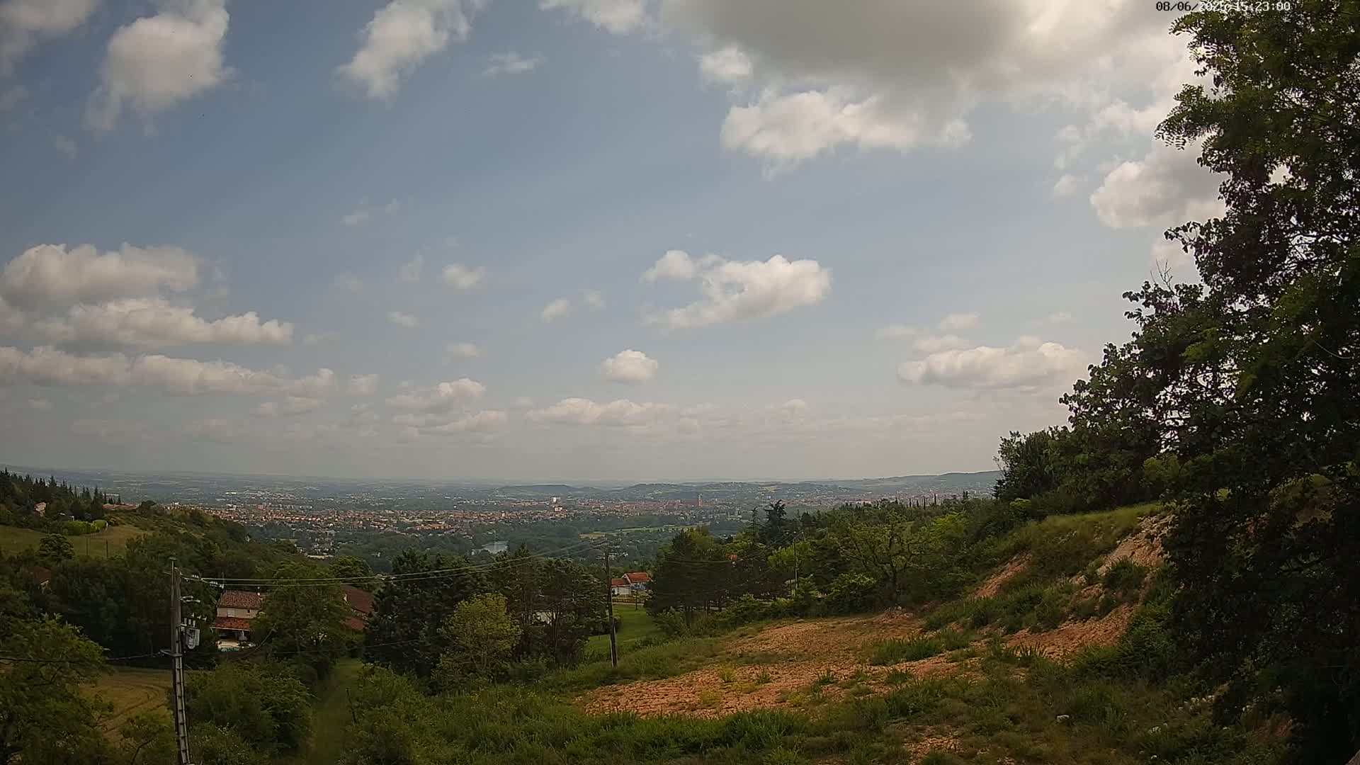 A partly cloudy day reveals a sprawling city in the distance, viewed from a hilltop with lush green vegetation and exposed, reddish-brown earth.
