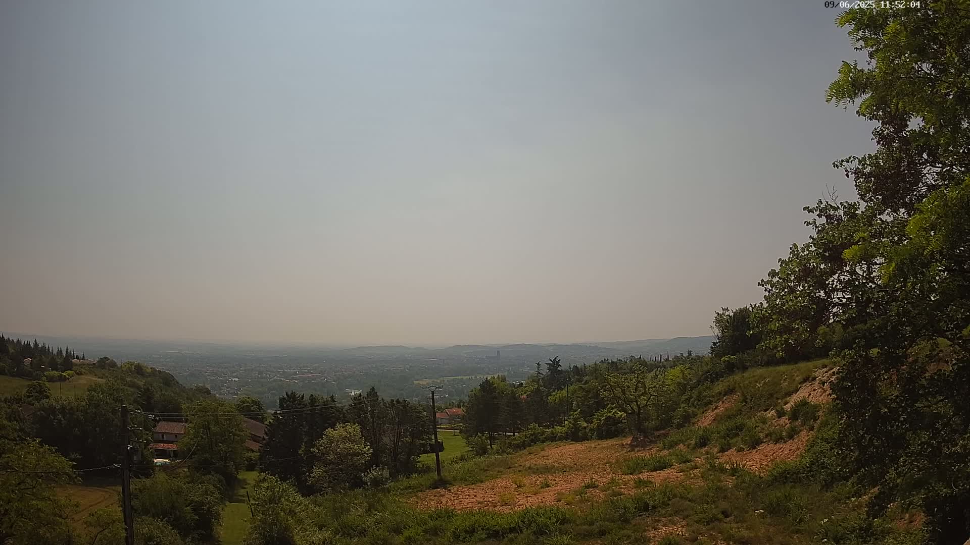 A hazy, sunny day reveals a panoramic view of a distant city from a hilltop, where lush green trees and shrubs surround a cleared, brown patch of land.