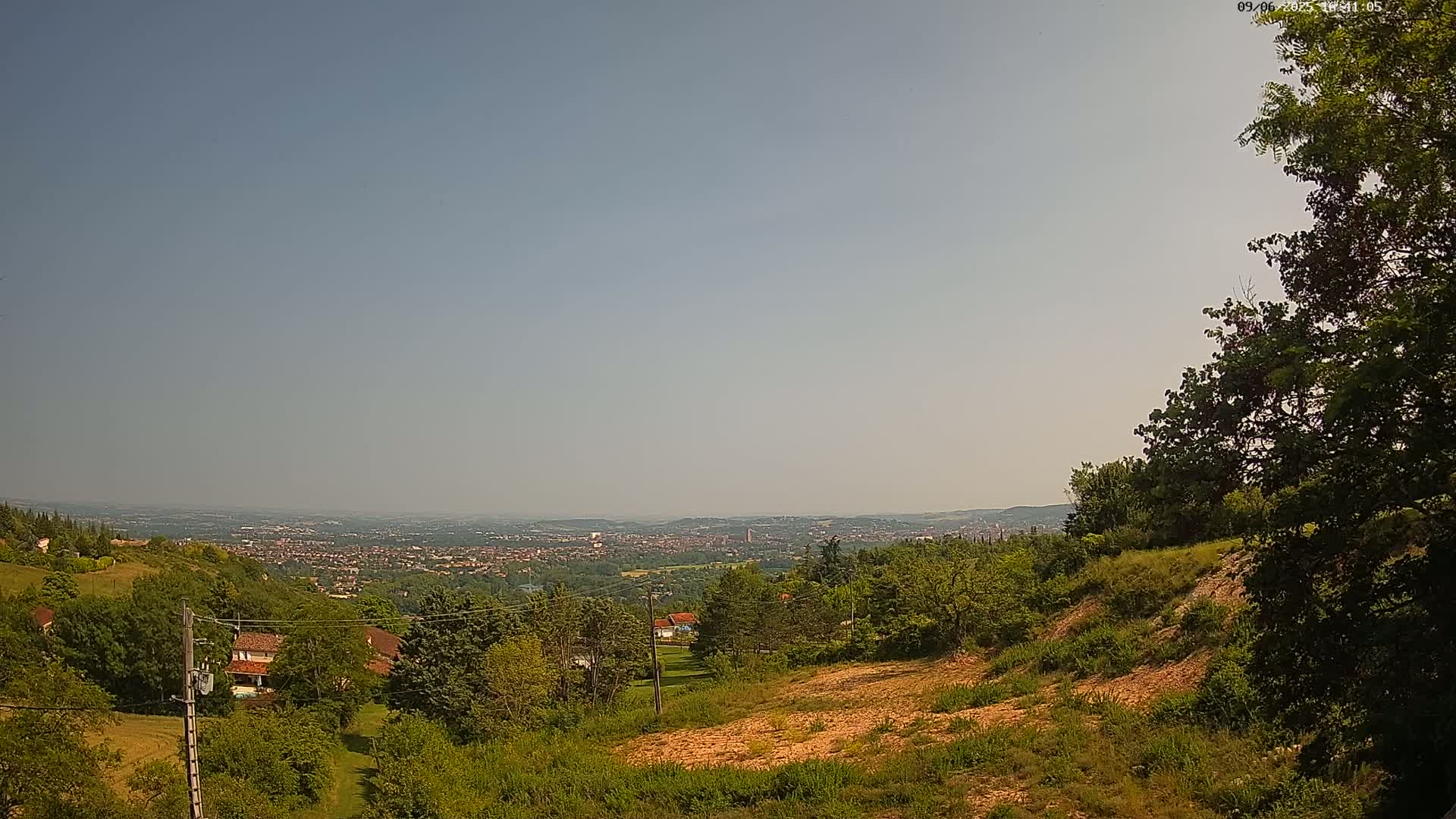 A hazy, sunny day reveals a sprawling city in the distance, viewed from a verdant hillside dotted with trees and houses.