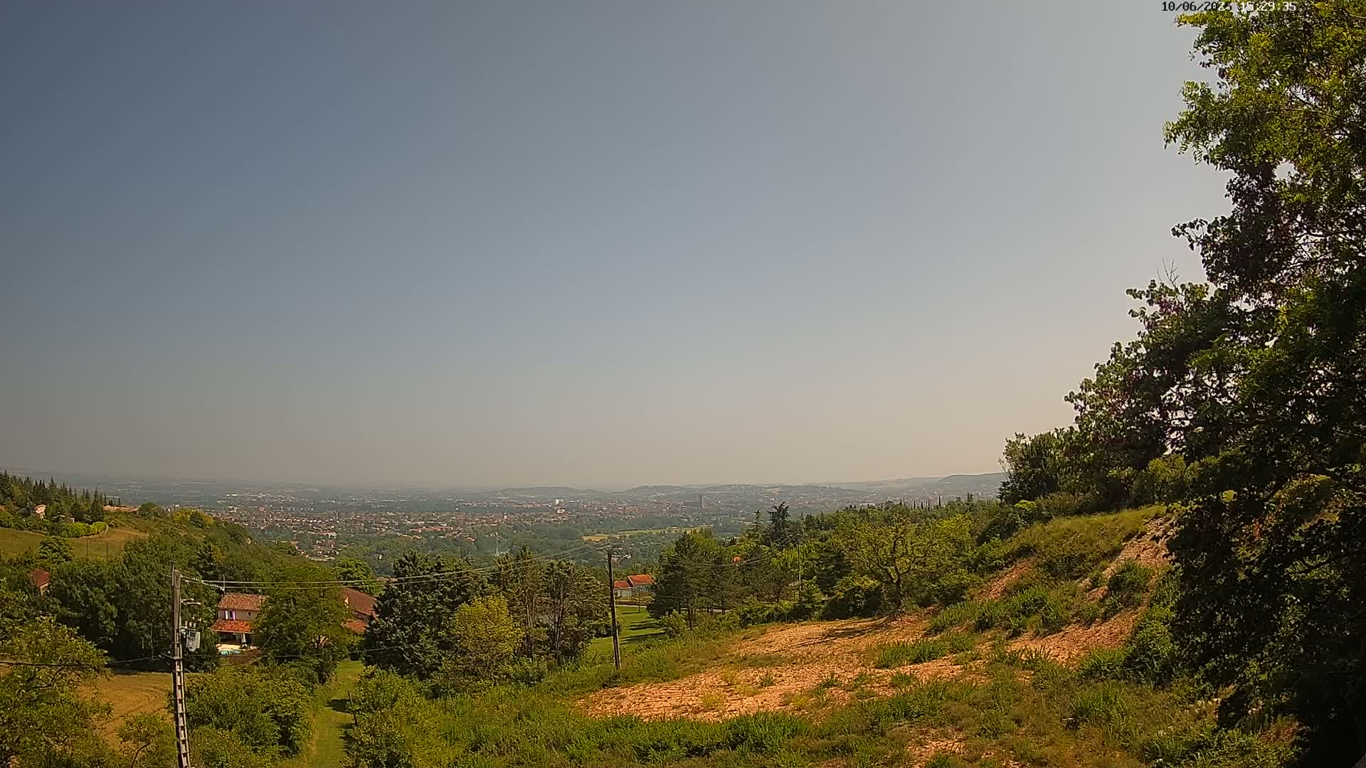 A hazy, sunny day reveals a panoramic view of a sprawling city nestled in a valley, seen from a high vantage point amidst lush green hills and trees.