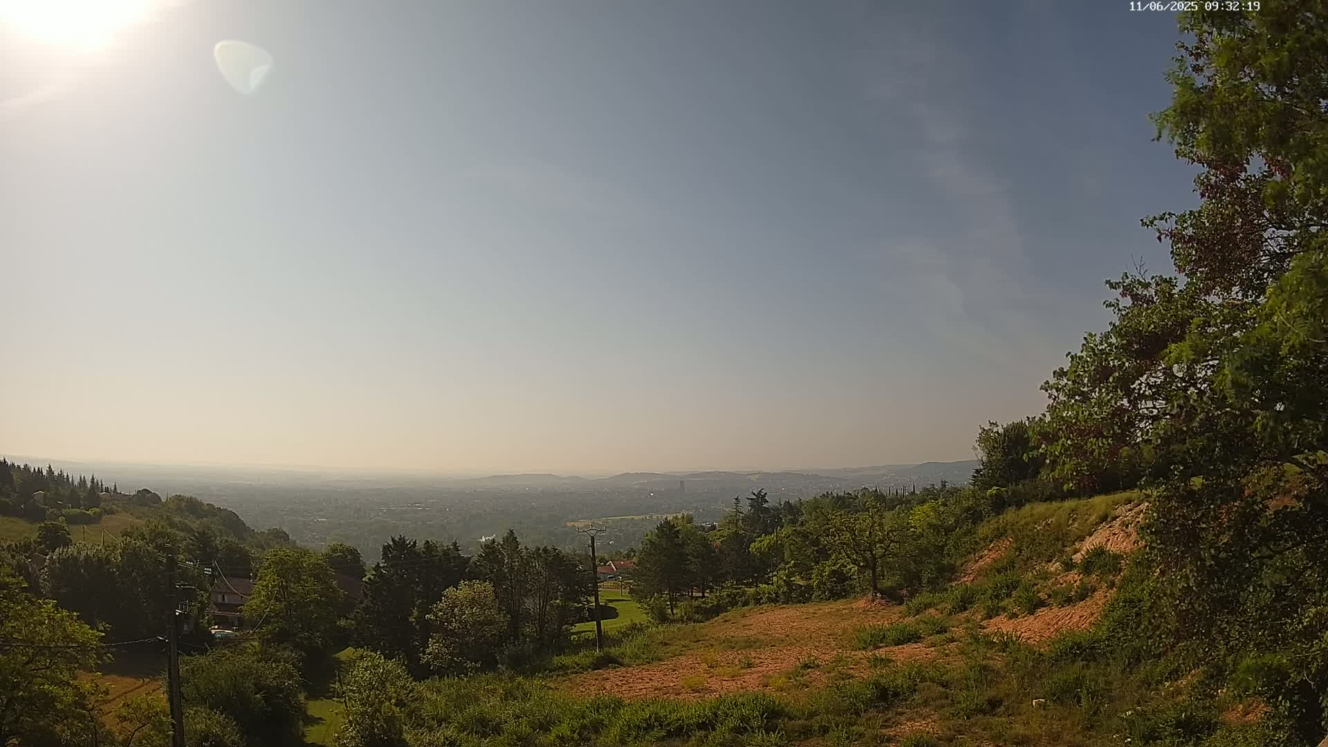 A hazy sunny day reveals a panoramic view of a sprawling city in the distance, nestled among rolling green hills and lush vegetation.
