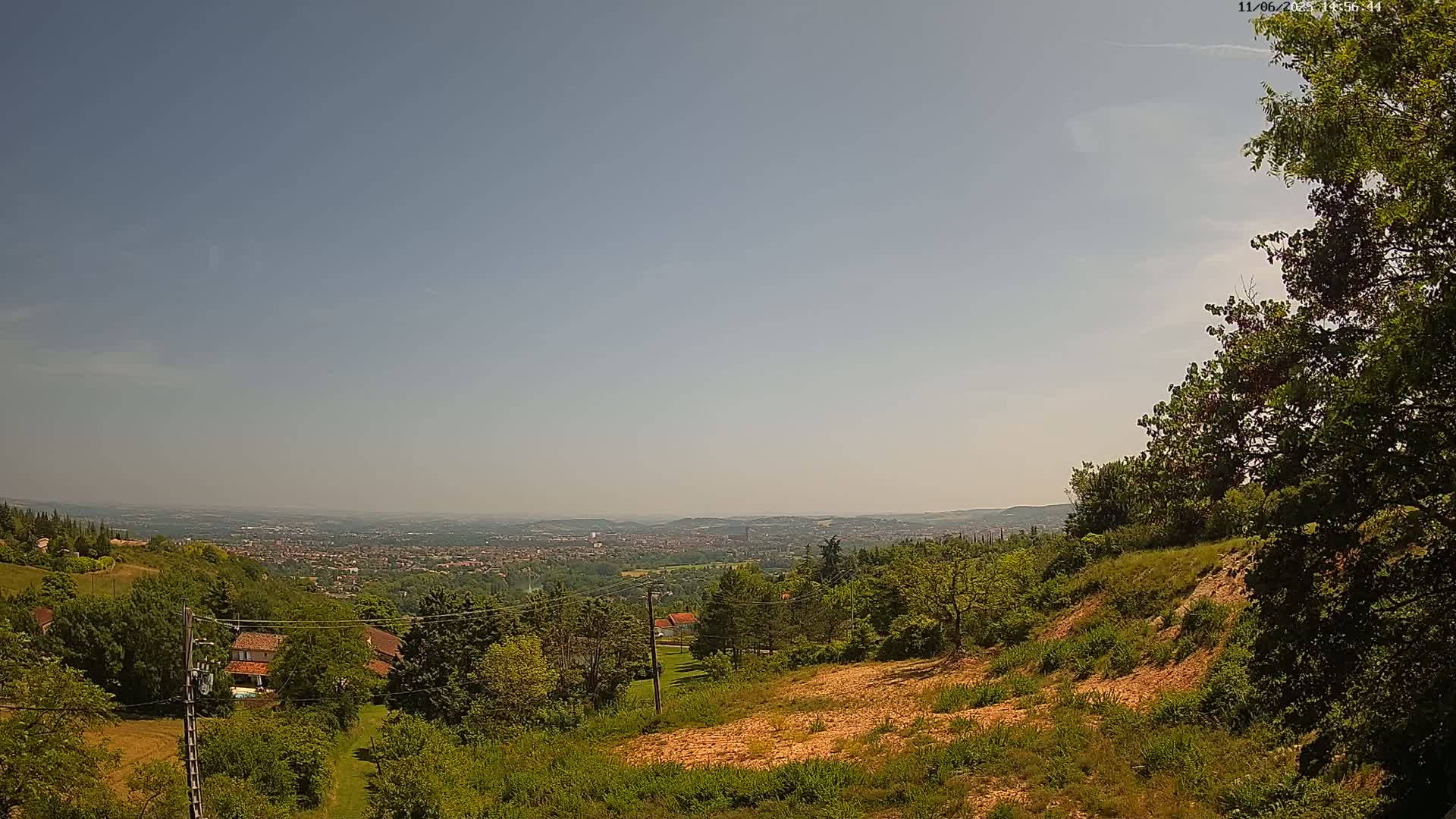 A hazy, sunny day reveals a sprawling view of a city nestled in a valley, seen from a hilltop covered in lush green vegetation and bare patches of earth.