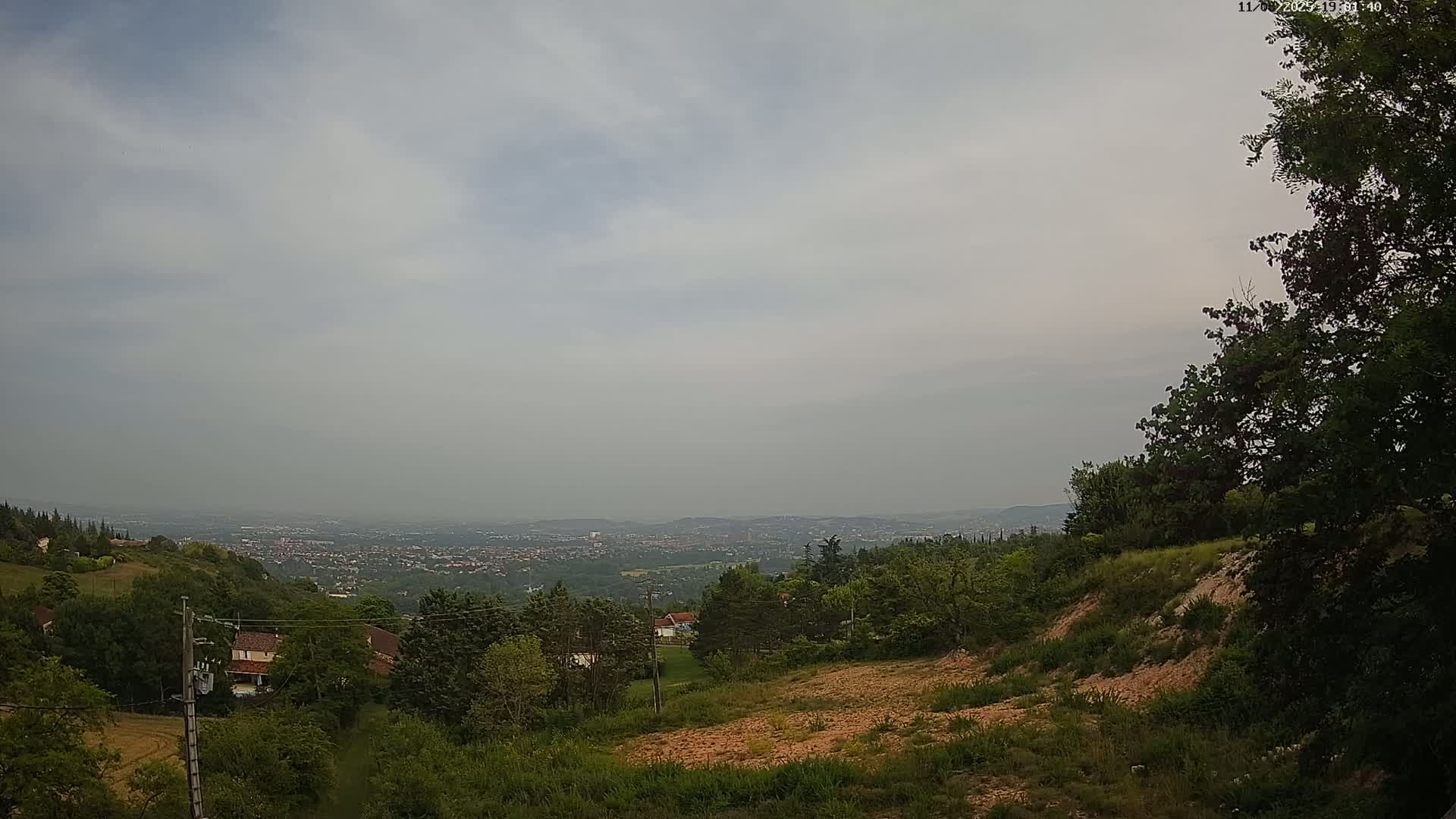 A hazy, overcast day reveals a hilly landscape with greenery, a distant city, and bare patches of earth.