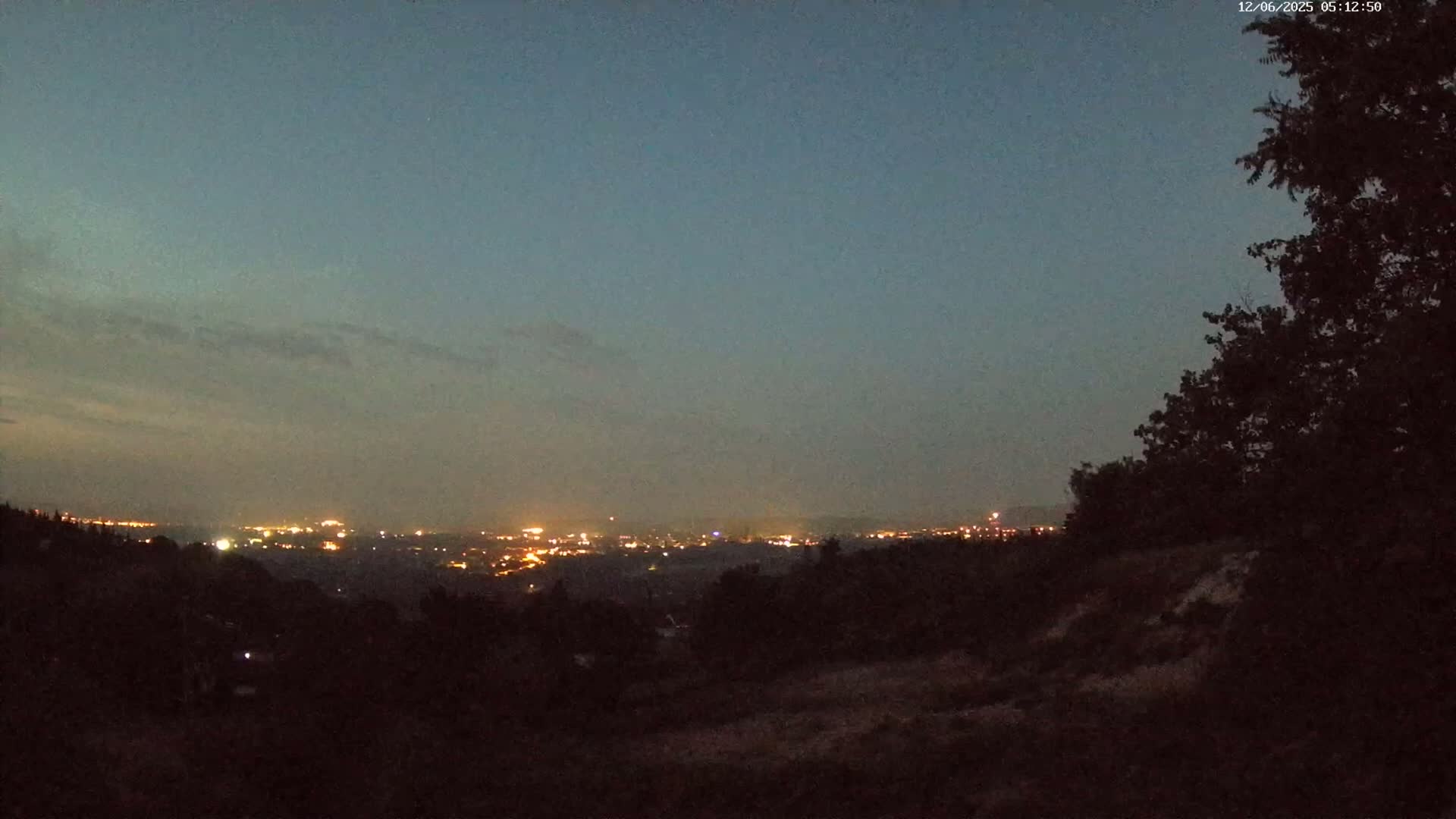 A distant city's lights illuminate the twilight sky, viewed from a hilltop amidst dark trees under a mostly clear, dusky sky.
