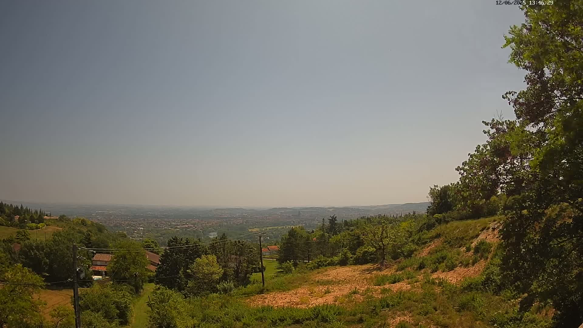 A hazy, sunny day reveals a panoramic view of a sprawling city nestled in a valley, overlooked by a lush, tree-covered hillside dotted with houses.
