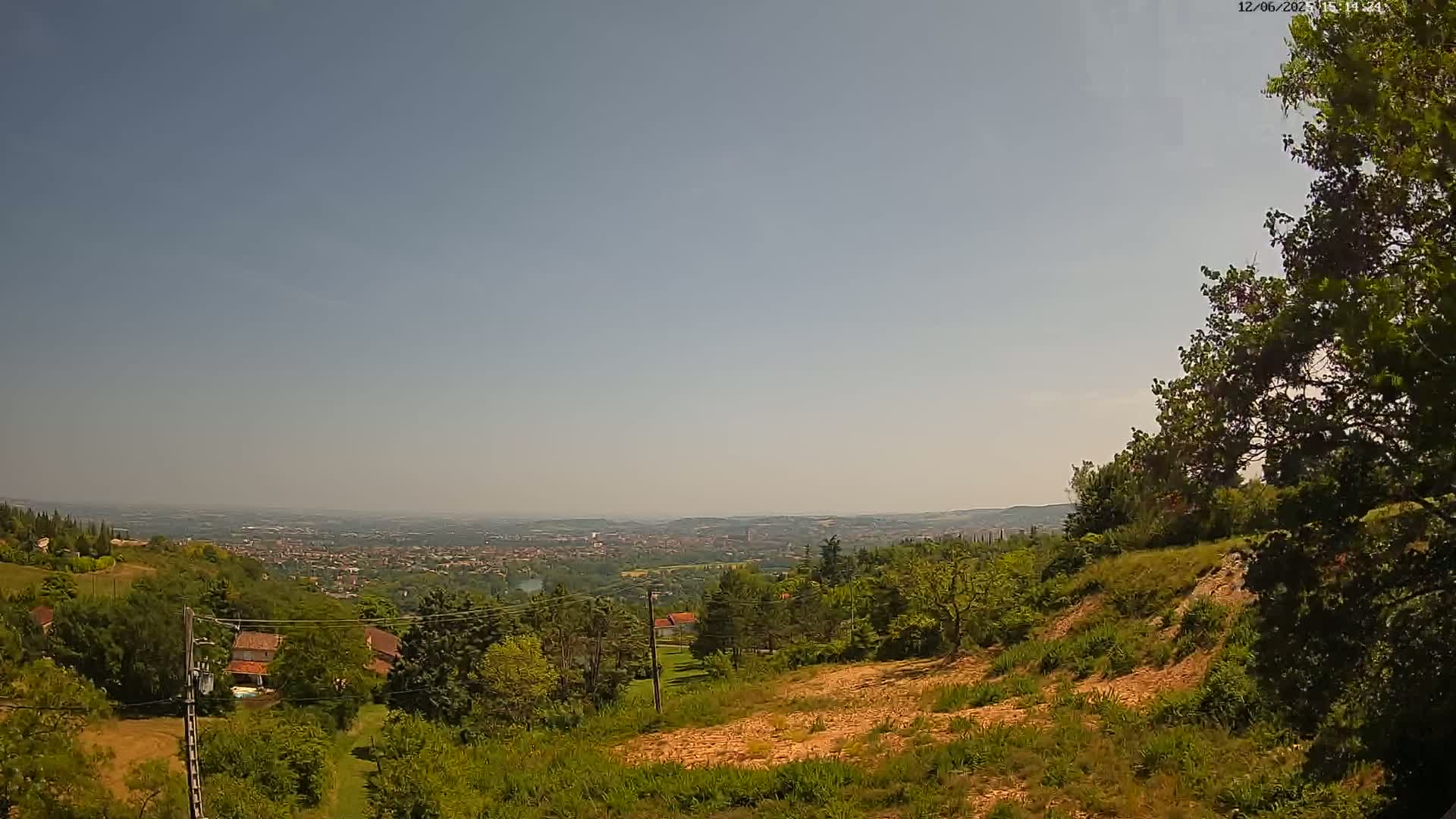 A hilly landscape under a clear, bright sky shows a distant city sprawling below, with lush green vegetation and several houses visible in the foreground.
