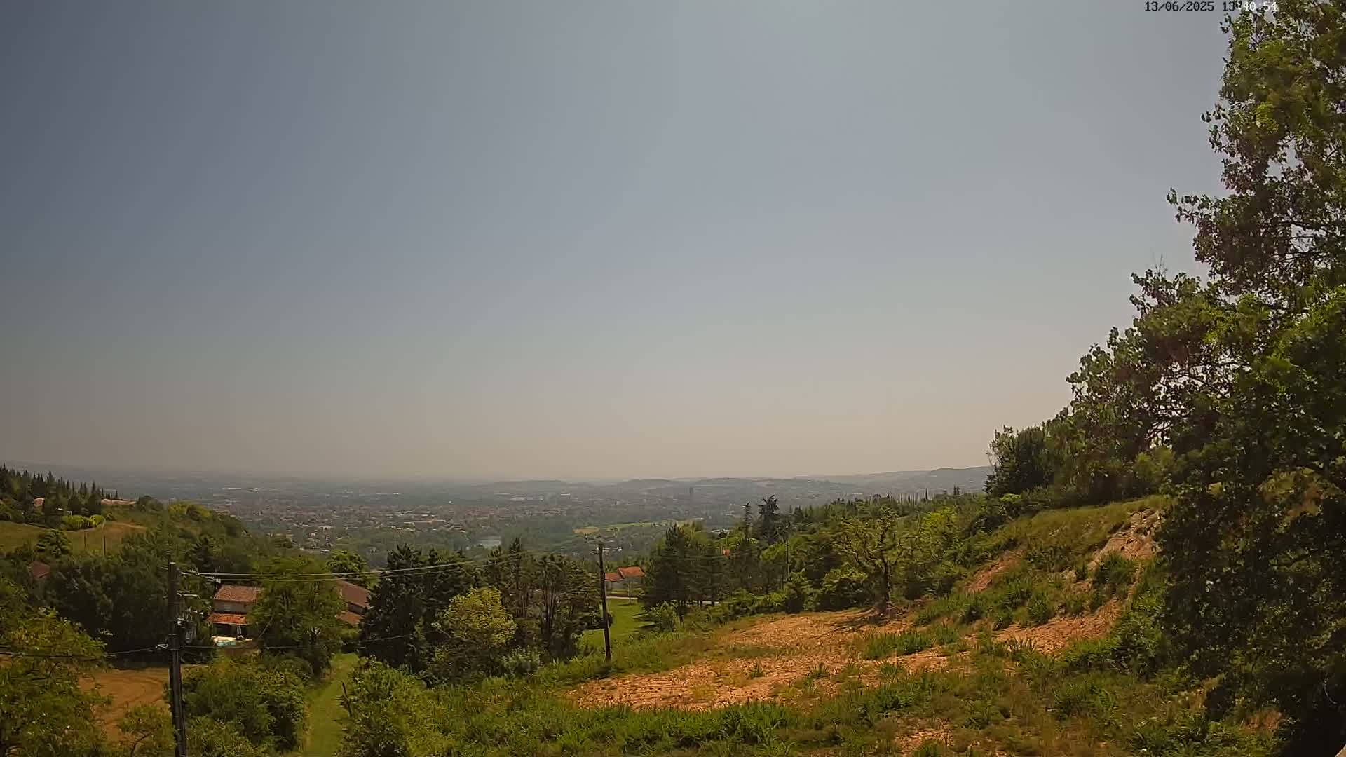 A hazy, sunny day reveals a sprawling vista of a distant city nestled in a valley, visible from a hilltop dotted with green trees and a few houses.