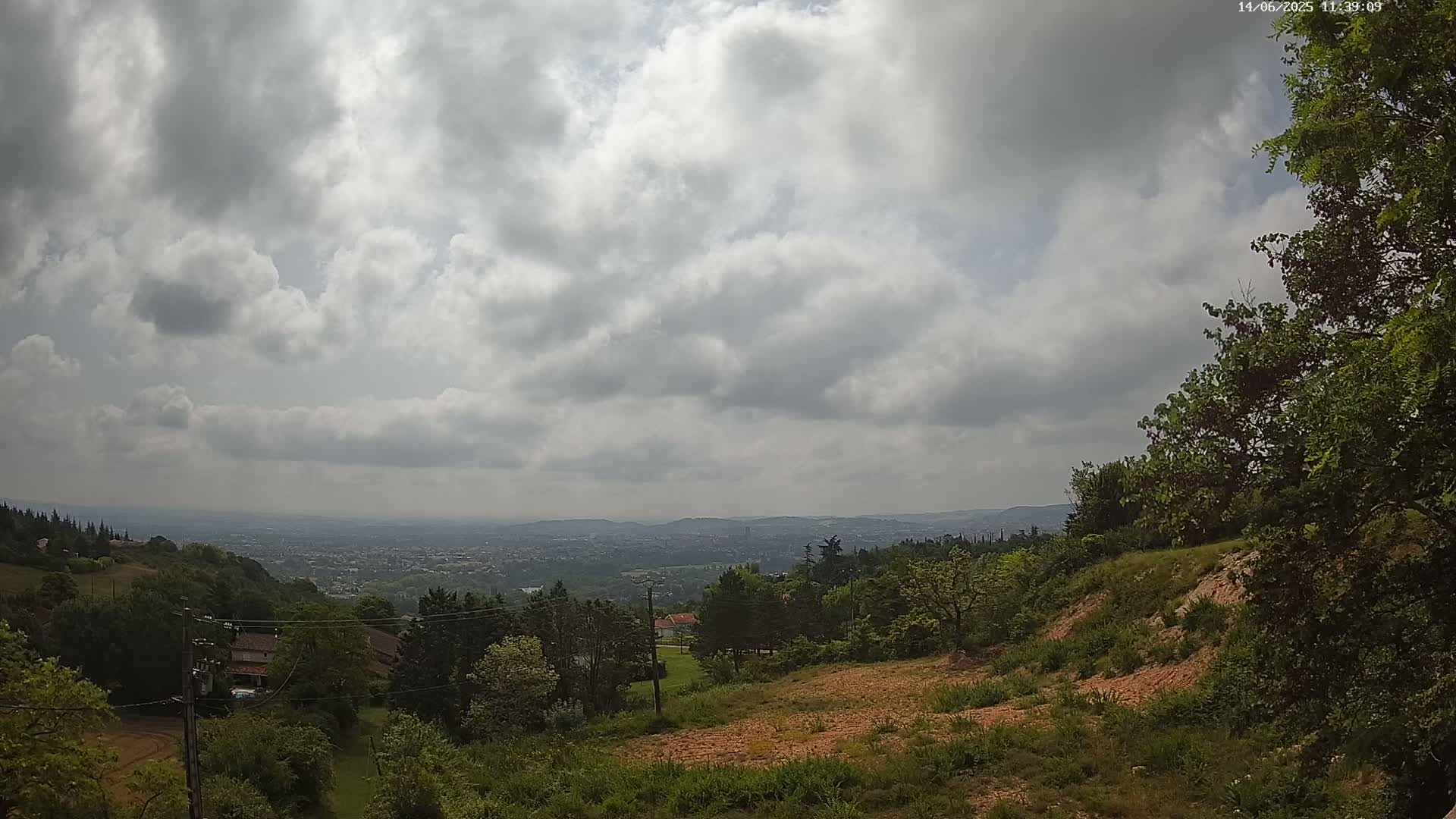 A partly cloudy sky overlooks a hilly landscape with green trees and a distant city under a hazy atmosphere.