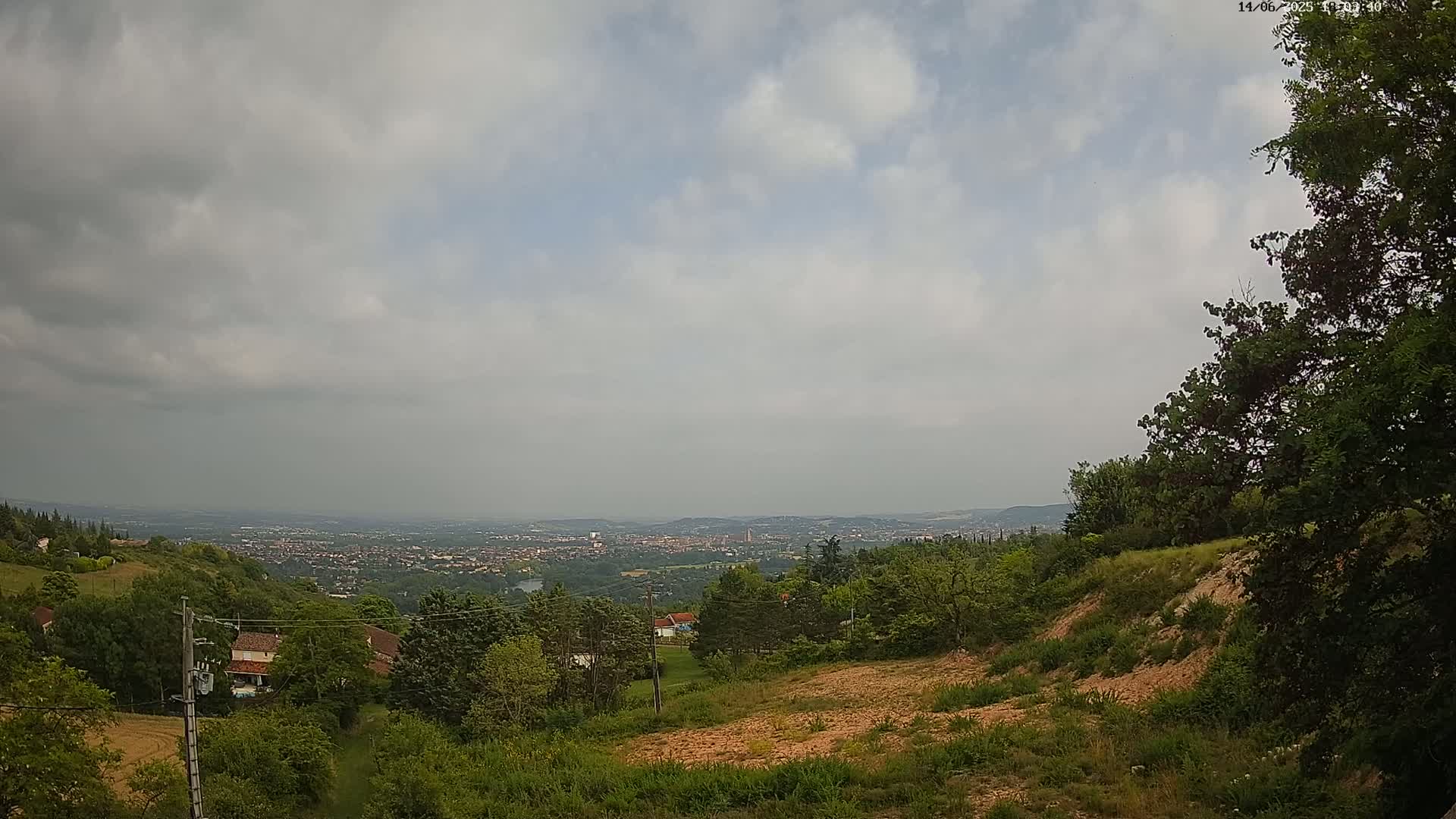 A hilly landscape with lush green vegetation overlooks a distant city under a cloudy sky.
