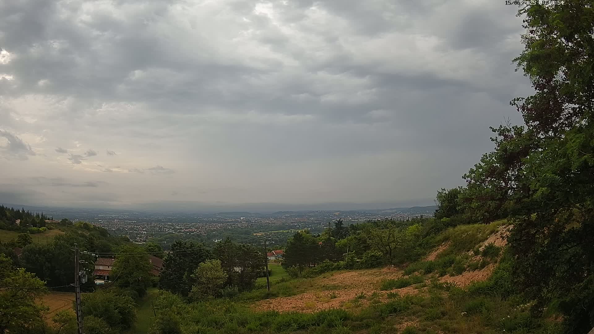 A hilly landscape under an overcast sky overlooks a distant city, with vegetation and a few houses visible in the foreground.