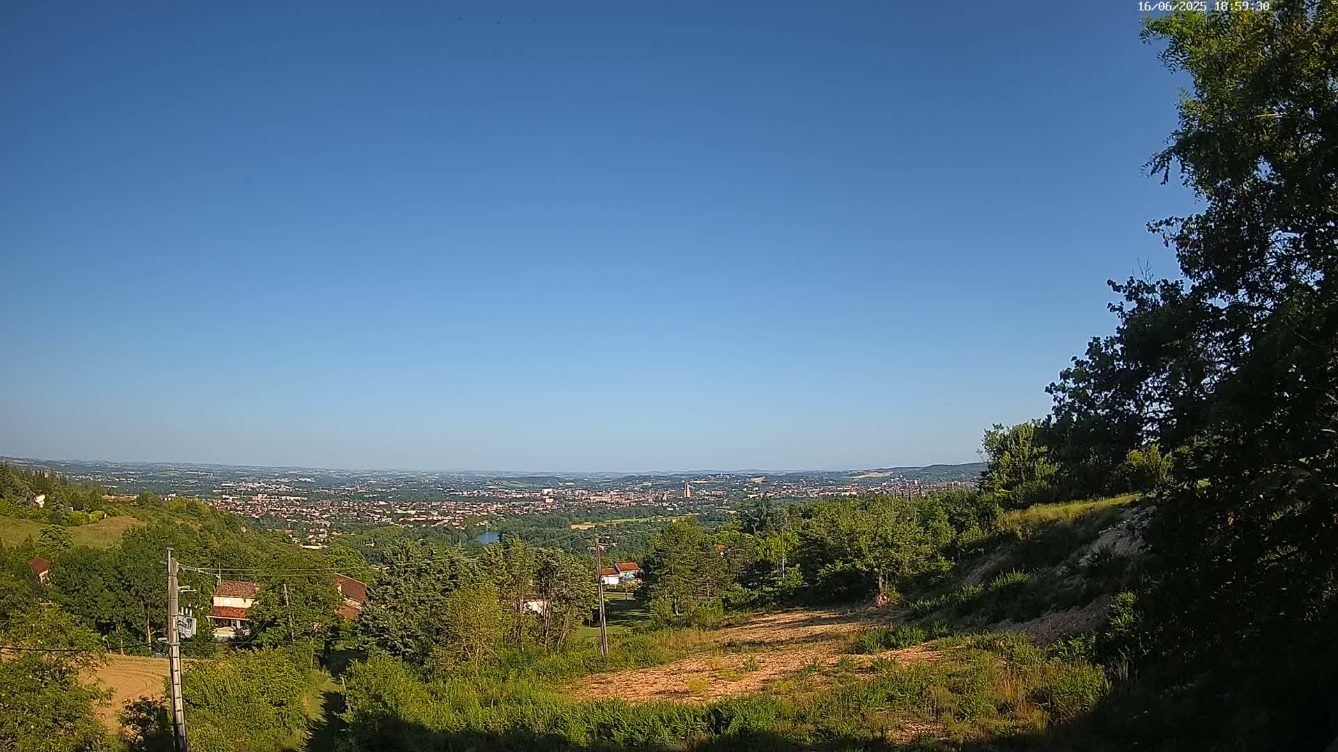 A sprawling city nestled in a valley is visible in the distance under a clear, blue sky, viewed from a hilltop vantage point with lush green vegetation in the foreground.