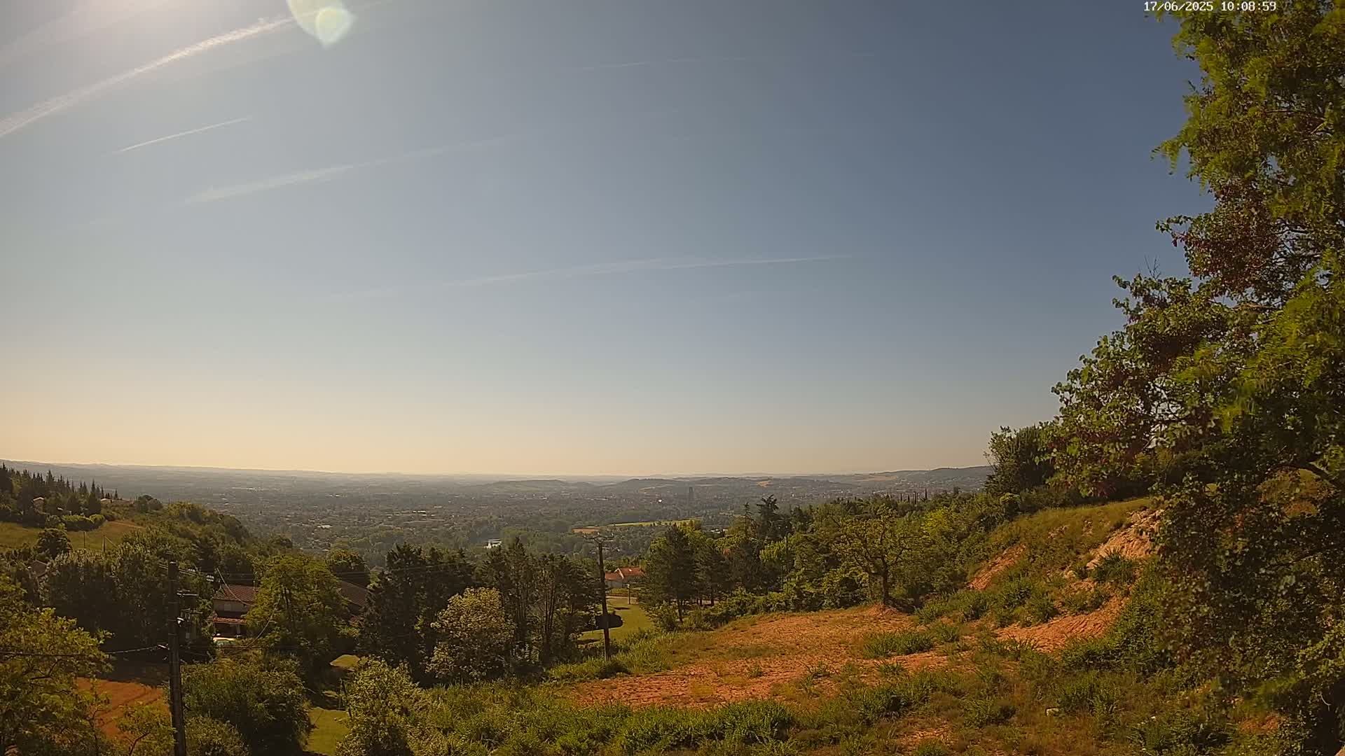 A sunny, clear day reveals a panoramic view of a distant city nestled in a valley, seen from a high vantage point amid a landscape of lush green trees and brown, grassy hills.