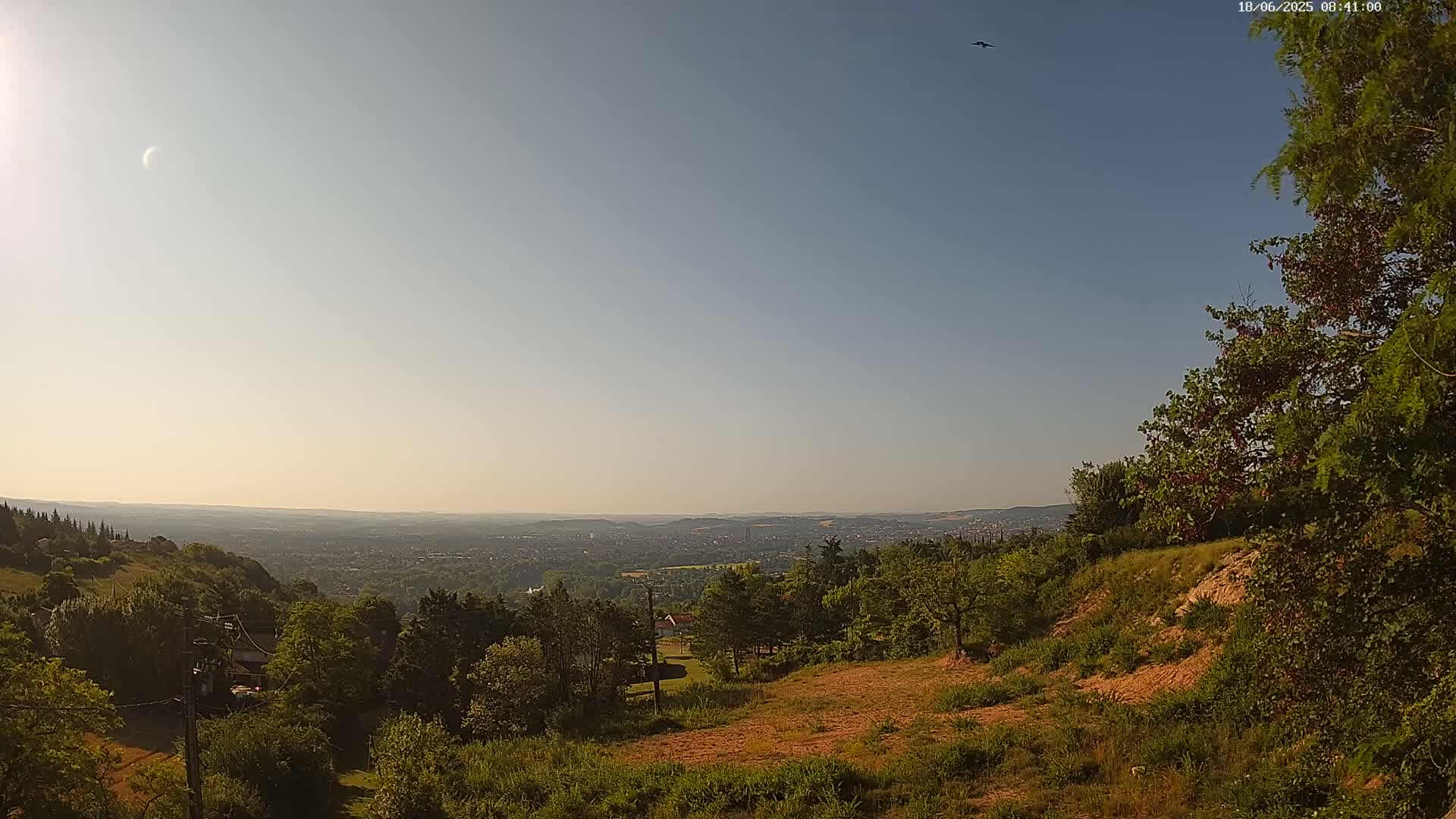 A sunny, clear day reveals a panoramic view of a sprawling city from a hilltop, surrounded by lush green trees and vegetation.