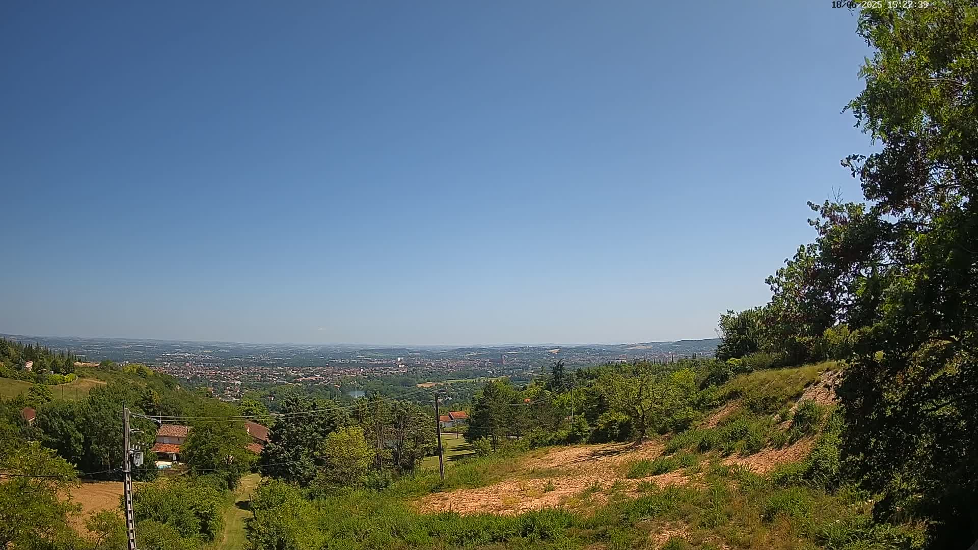A sprawling vista of a city in the distance, under a clear blue sky, is visible from a verdant hillside with houses and trees in the foreground.