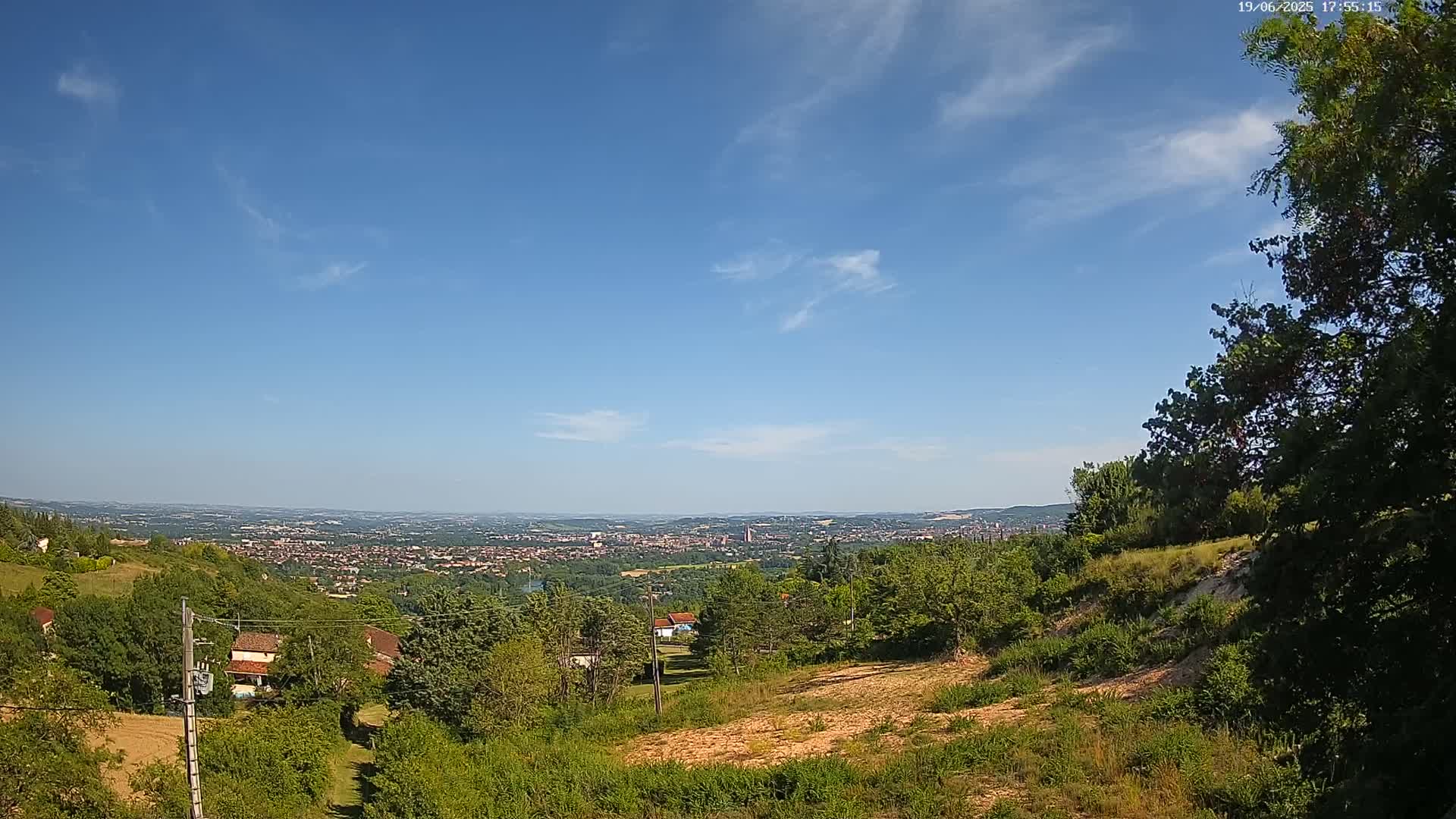 A sprawling city is visible in the distance under a mostly clear blue sky, viewed from a hilltop covered in green vegetation.
