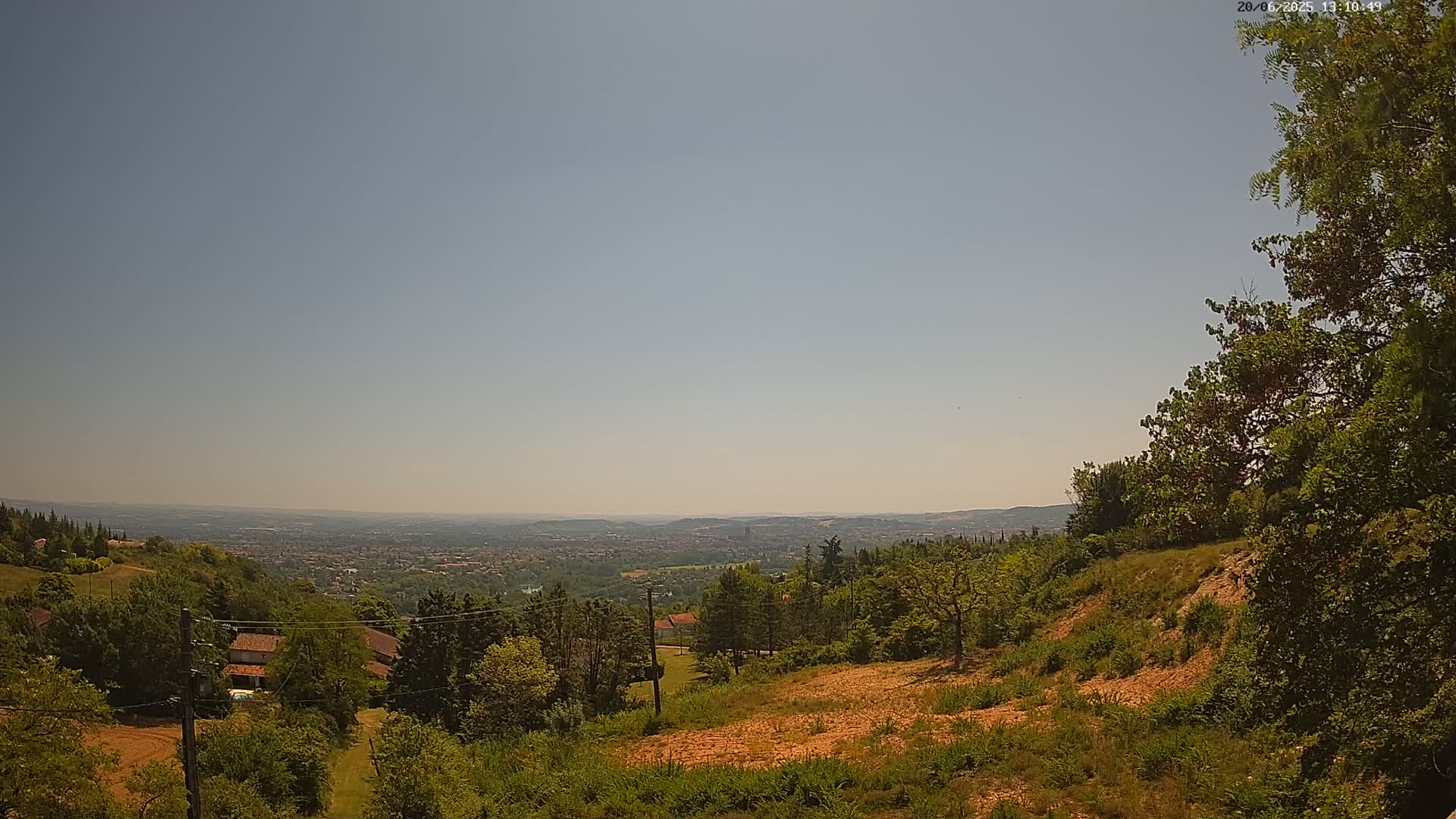 A hazy, sunny day reveals a sprawling vista of a city nestled in a valley, viewed from a tree-covered hillside with a few houses and bare patches of earth visible in the foreground.