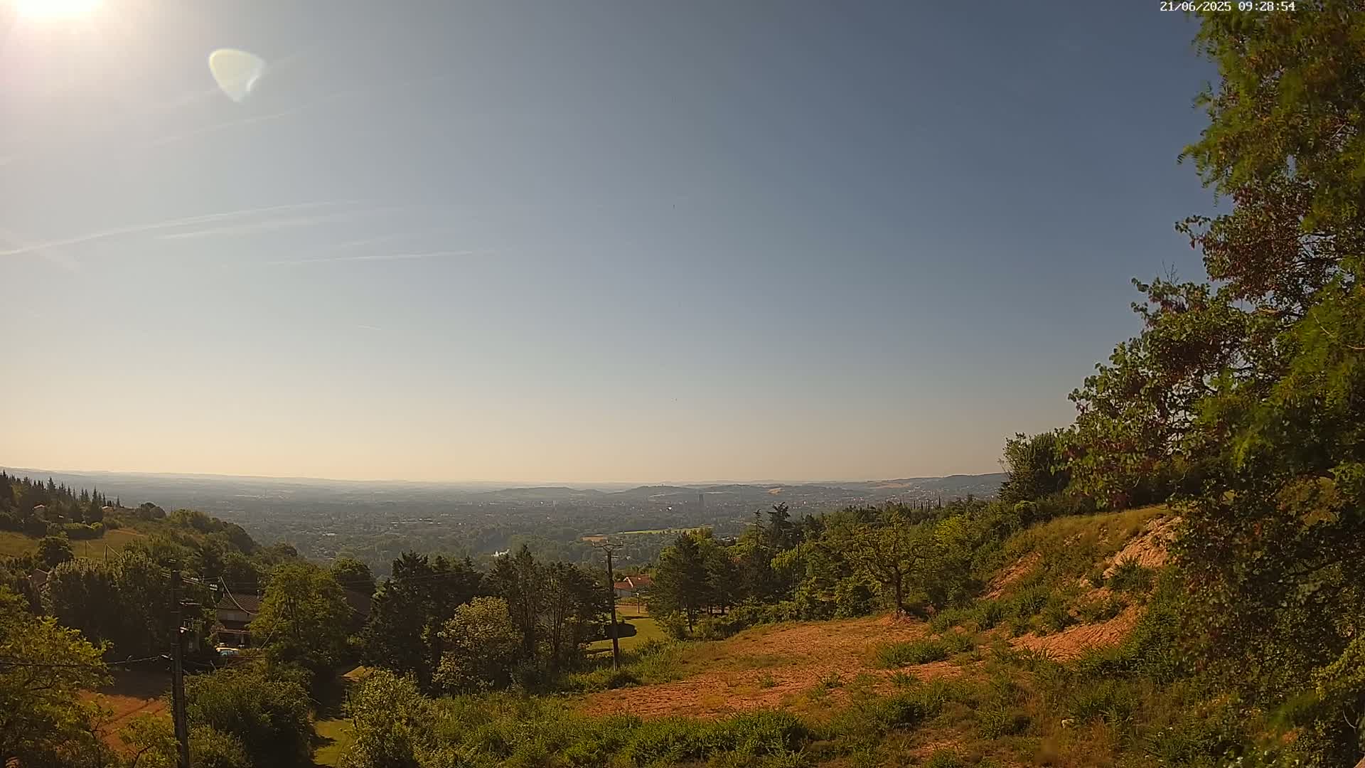 A sunny, hazy day reveals a panoramic view of a sprawling city nestled in a valley, surrounded by lush green hills and forests.