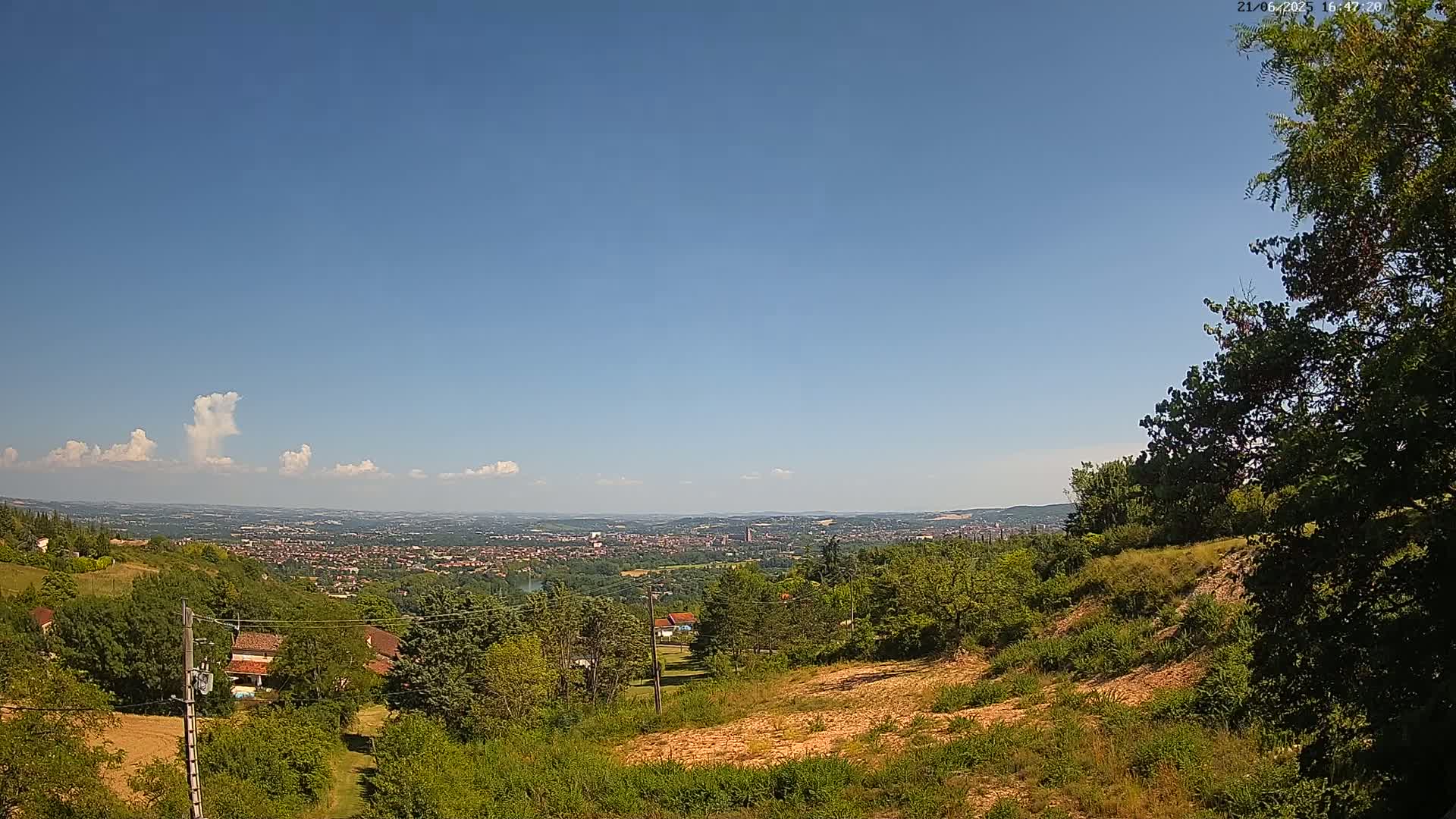 A hilly landscape with lush green vegetation overlooks a distant city under a mostly clear blue sky with a few scattered cumulus clouds.