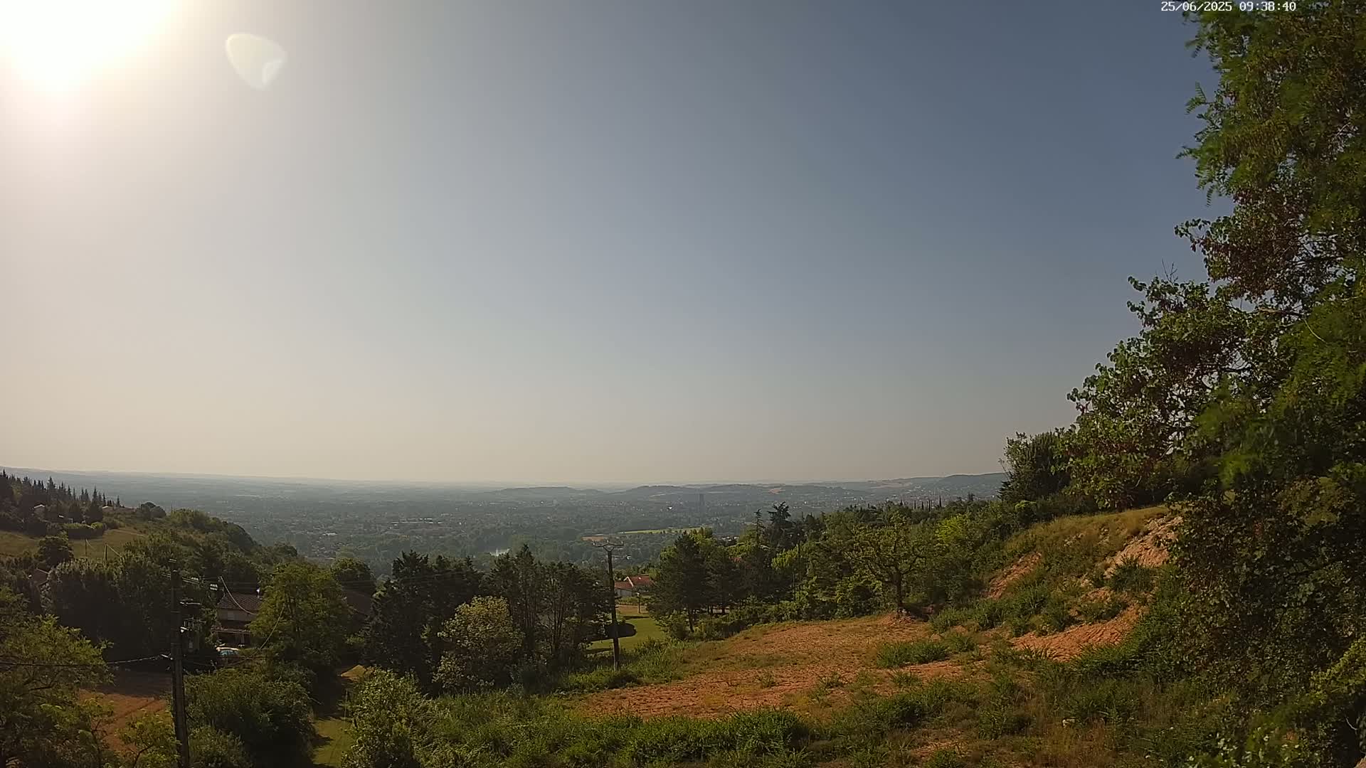 A sunny, hazy day reveals a panoramic view of a sprawling city nestled in a valley below a hillside covered in lush green trees and shrubs.