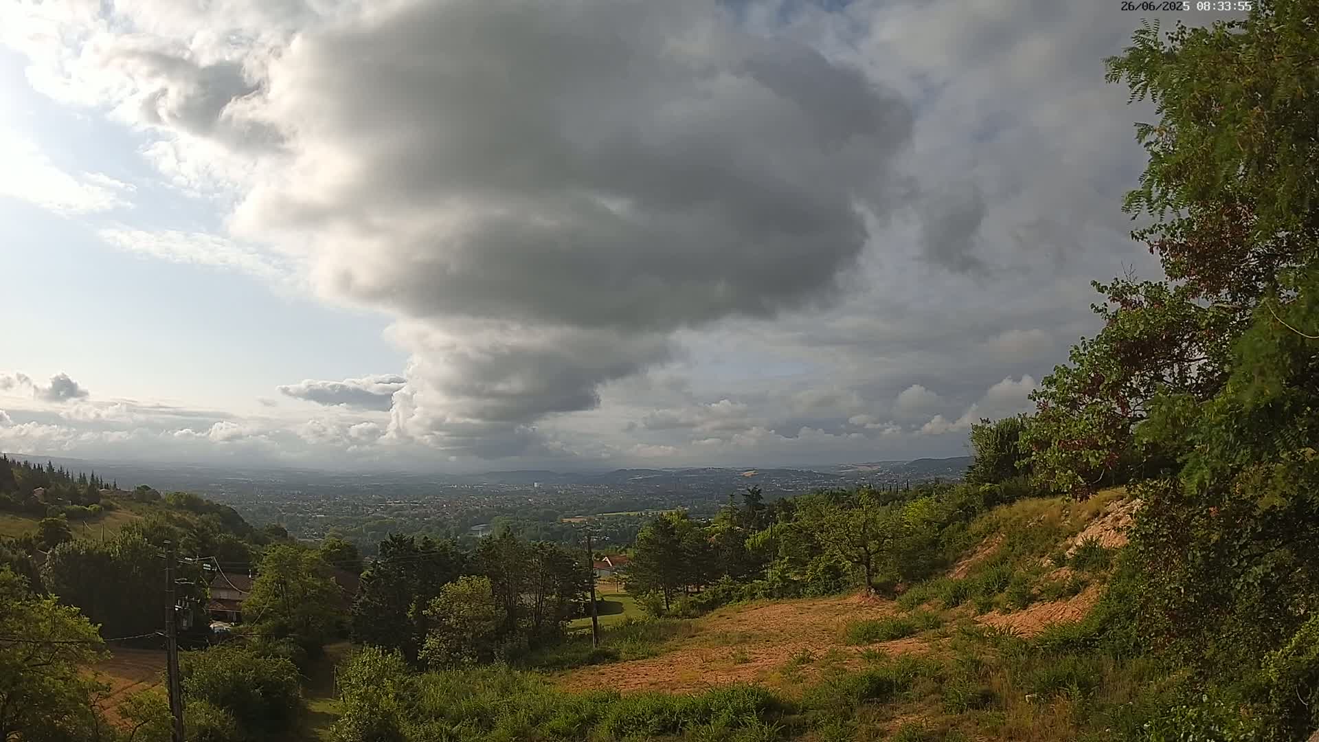 A partly cloudy sky hangs over a hilly landscape of green trees and shrubs, revealing a distant city in the valley below.