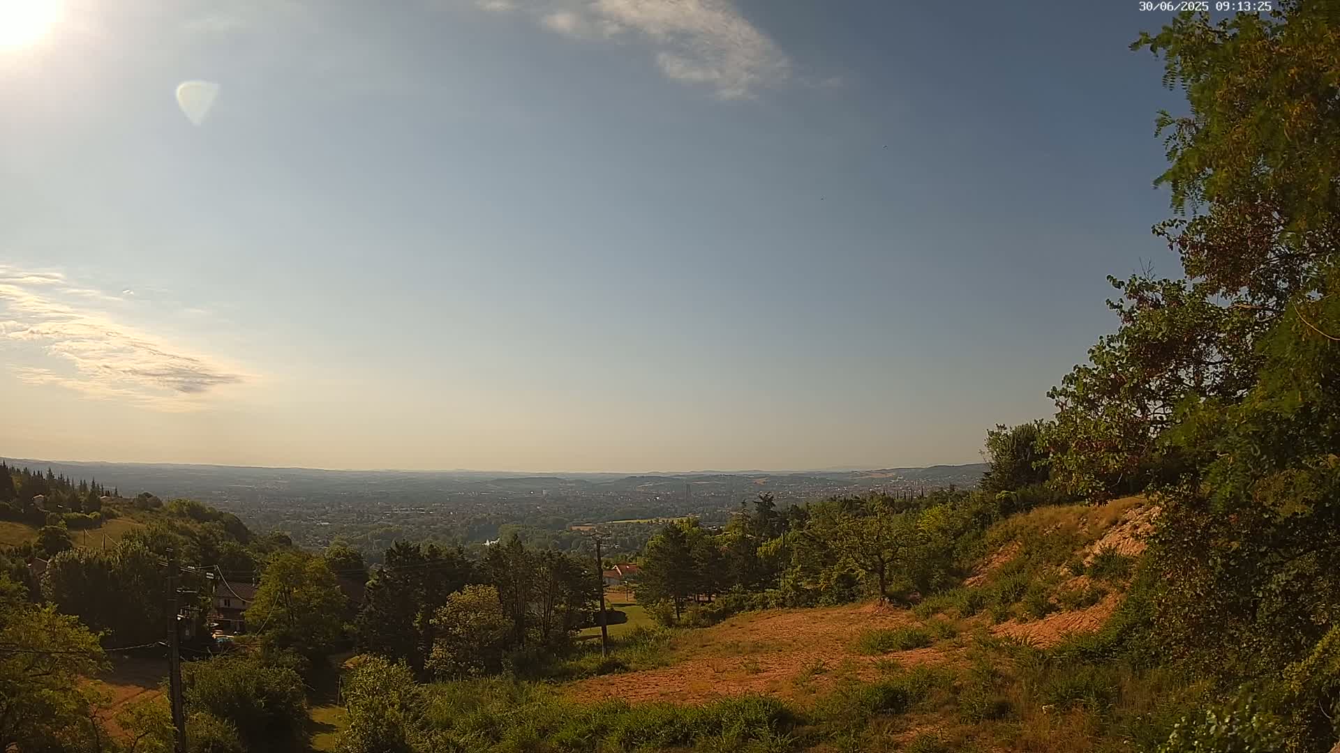A sunny day reveals a panoramic view of a distant city nestled within a valley, surrounded by lush green hills and trees.