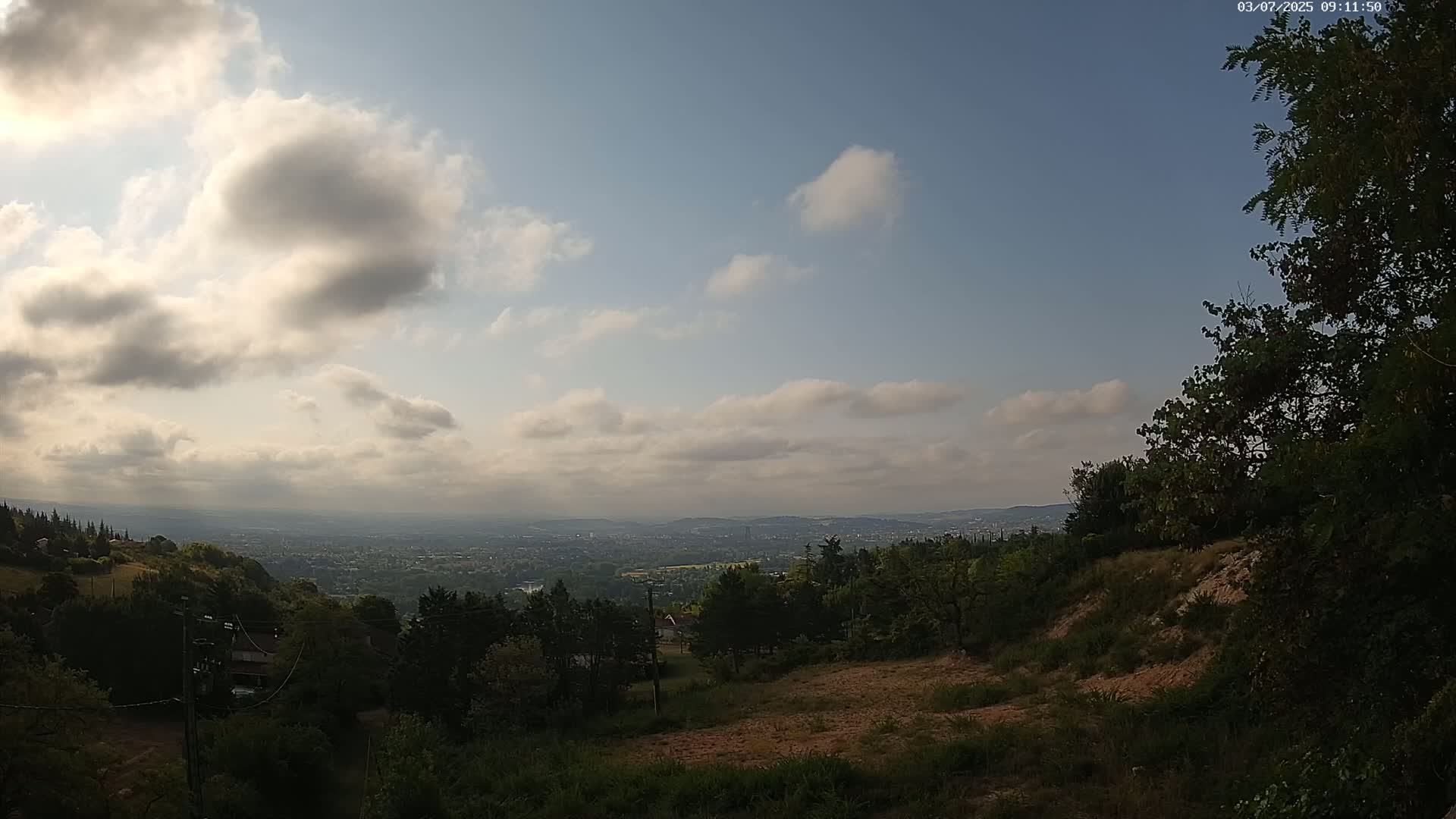 A partly cloudy sky overlooks a hilly landscape with a distant city visible below, under a hazy, sunny sky.