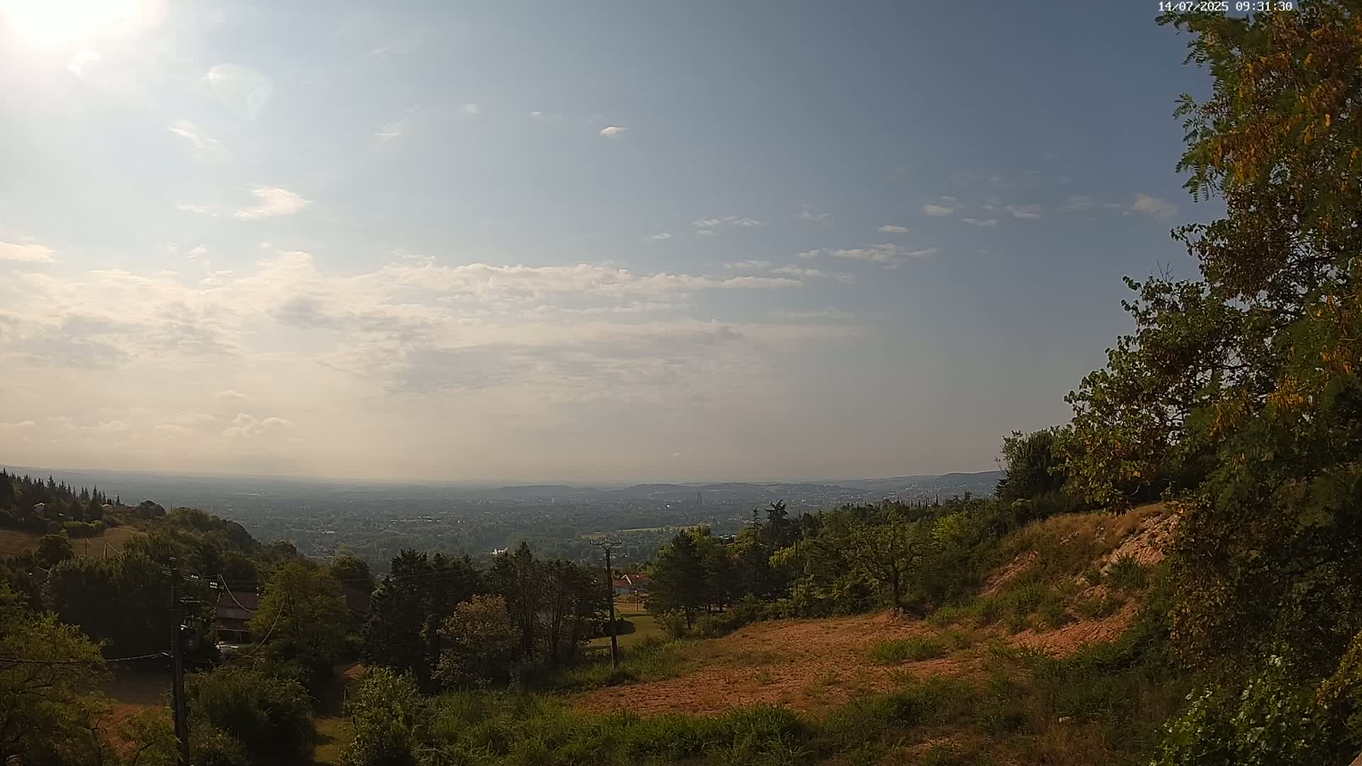 A partly sunny day reveals a view of a distant city sprawling across a valley, seen from a hilltop dotted with green trees and patches of brown earth.