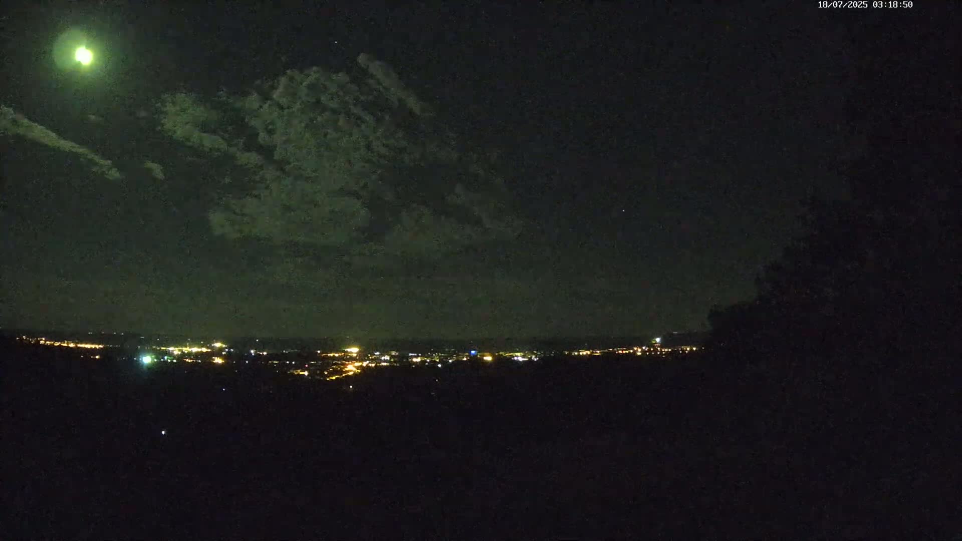A bright moon shines through thin clouds over a dark, distant cityscape at night.