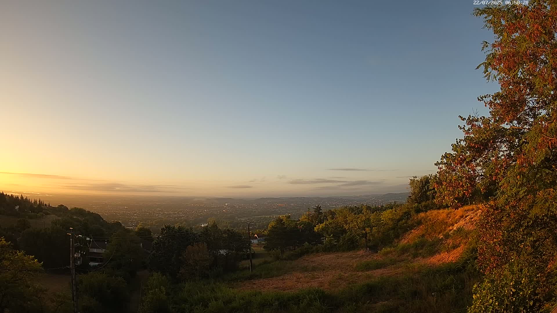 A hazy sunrise illuminates a sprawling cityscape viewed from a hilly, tree-covered vantage point.