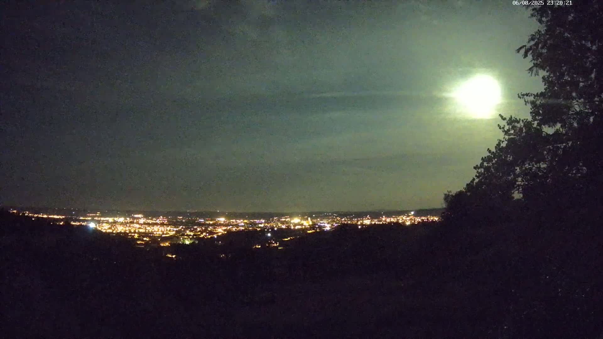 A brightly lit city is visible in the distance under a mostly cloudy night sky, illuminated by a large, bright moon partially obscured by clouds, as seen from an elevated vantage point with silhouetted trees in the foreground.