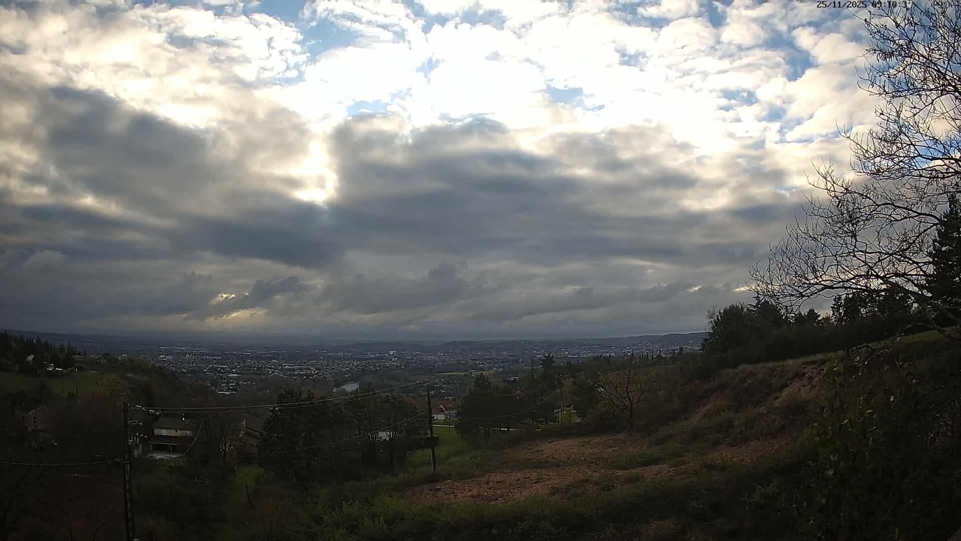 A panoramic outdoor view reveals a sprawling city in the distance under a dramatic, partly cloudy sky with bright sunlight breaking through dark clouds, framed by a hilly foreground dotted with bare trees and a few structures.
