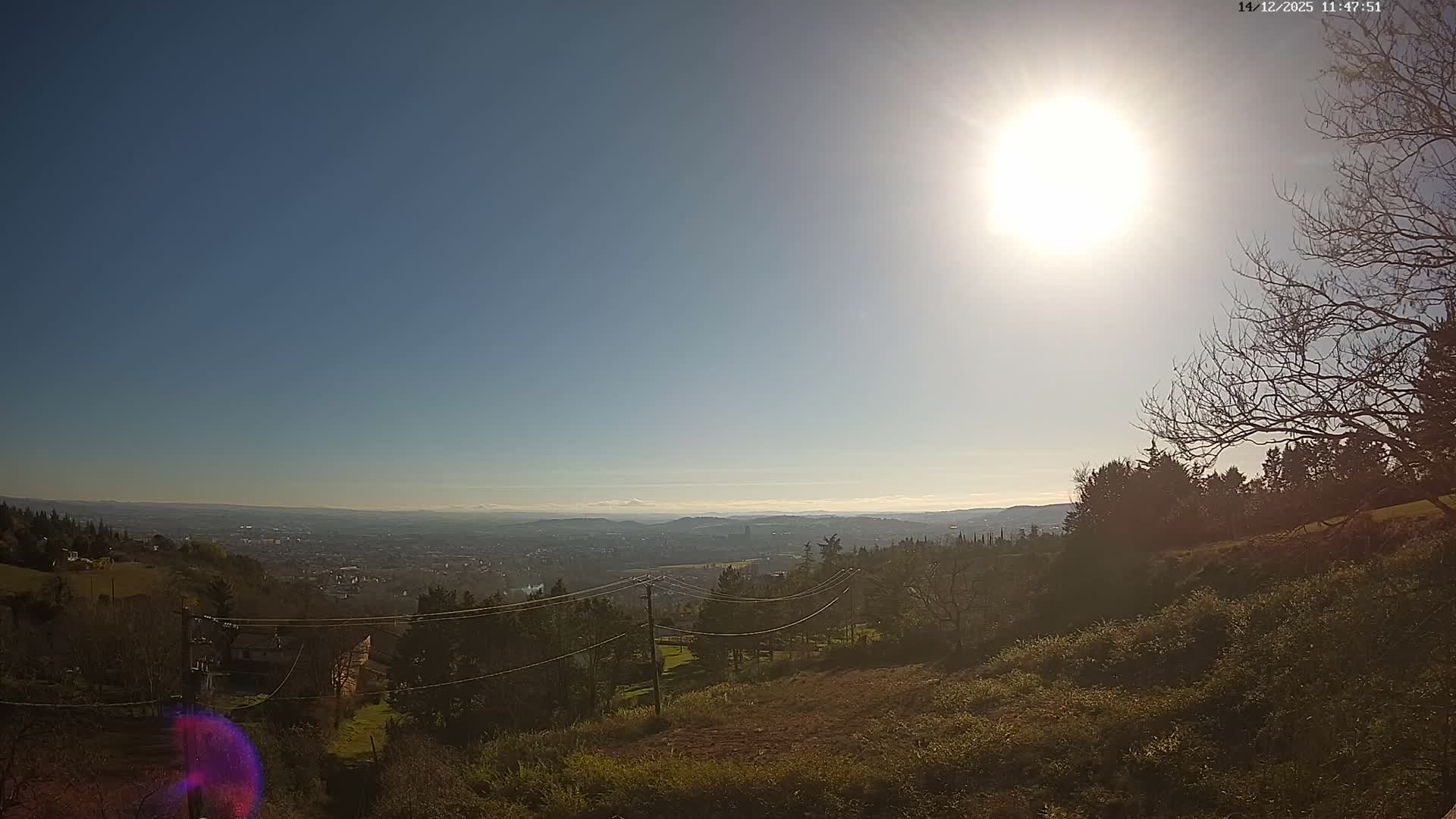 A panoramic outdoor view reveals a sprawling city in the distance under a dramatic, partly cloudy sky with bright sunlight breaking through dark clouds, framed by a hilly foreground dotted with bare trees and a few structures.