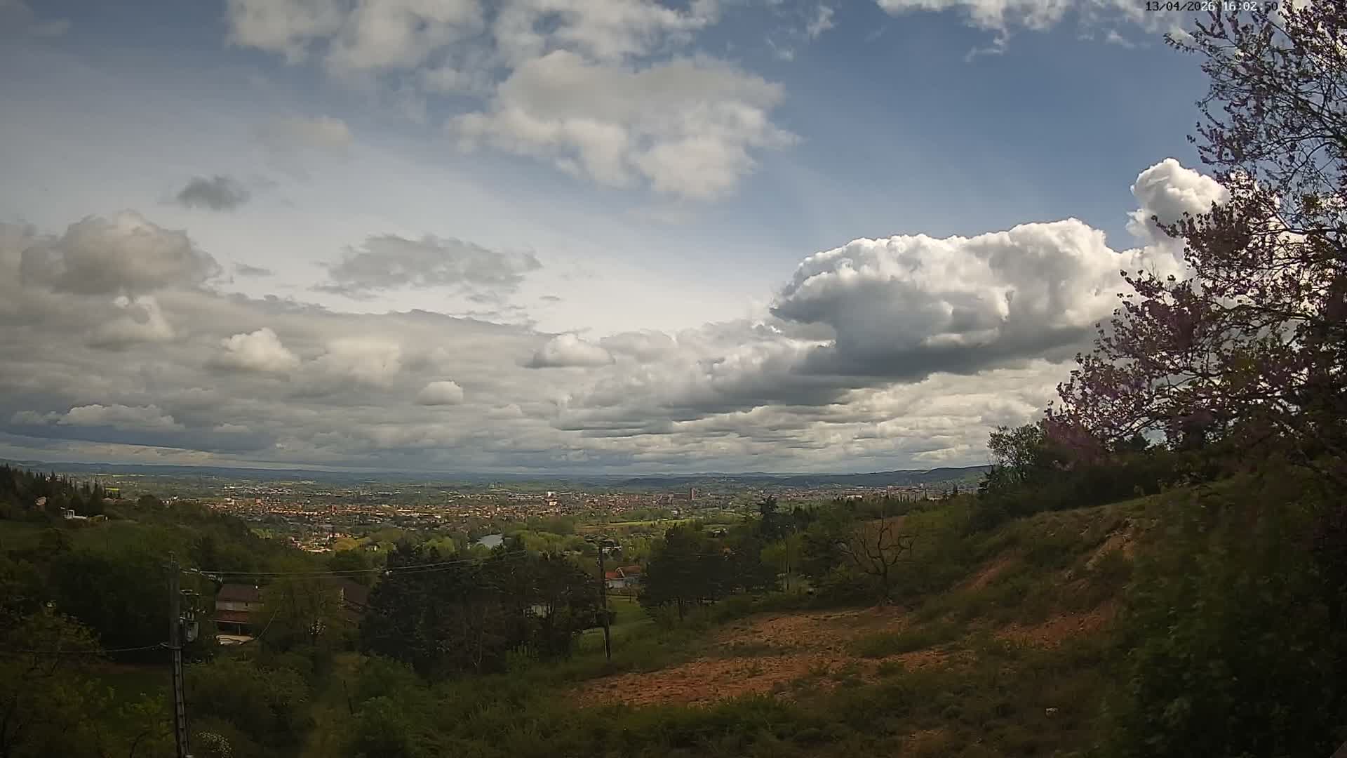 A panoramic outdoor view reveals a sprawling city in the distance under a dramatic, partly cloudy sky with bright sunlight breaking through dark clouds, framed by a hilly foreground dotted with bare trees and a few structures.