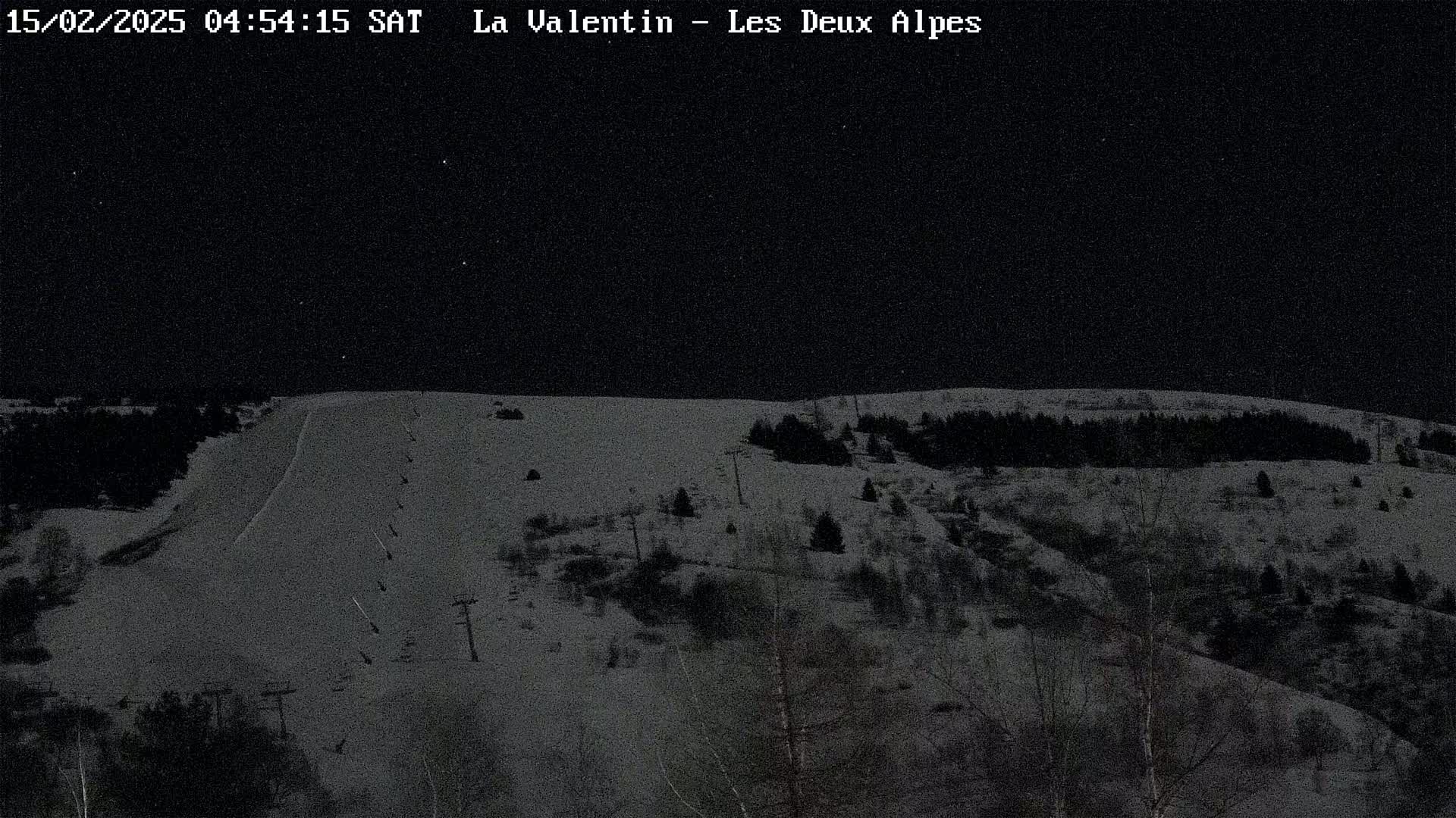 A snow-covered ski slope at night under a dark, starry sky, with sparsely scattered trees visible on the surrounding hills.