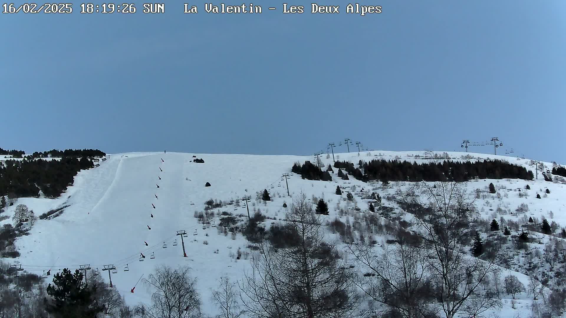 A snow-covered ski slope with several chairlifts and sparsely wooded areas under a clear, light blue sky.