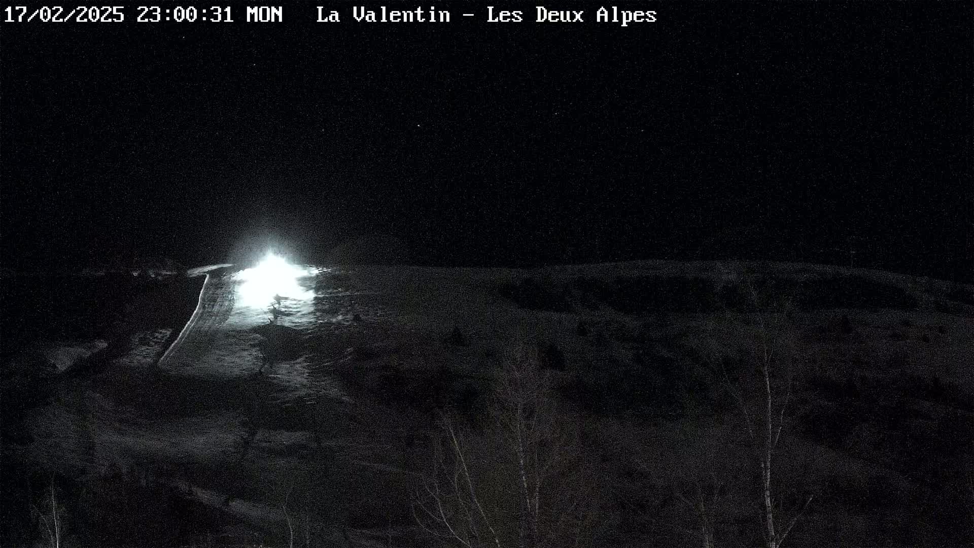A nighttime, low-light view of a snow-covered slope with a bright light source at the center, possibly from a vehicle's headlights.