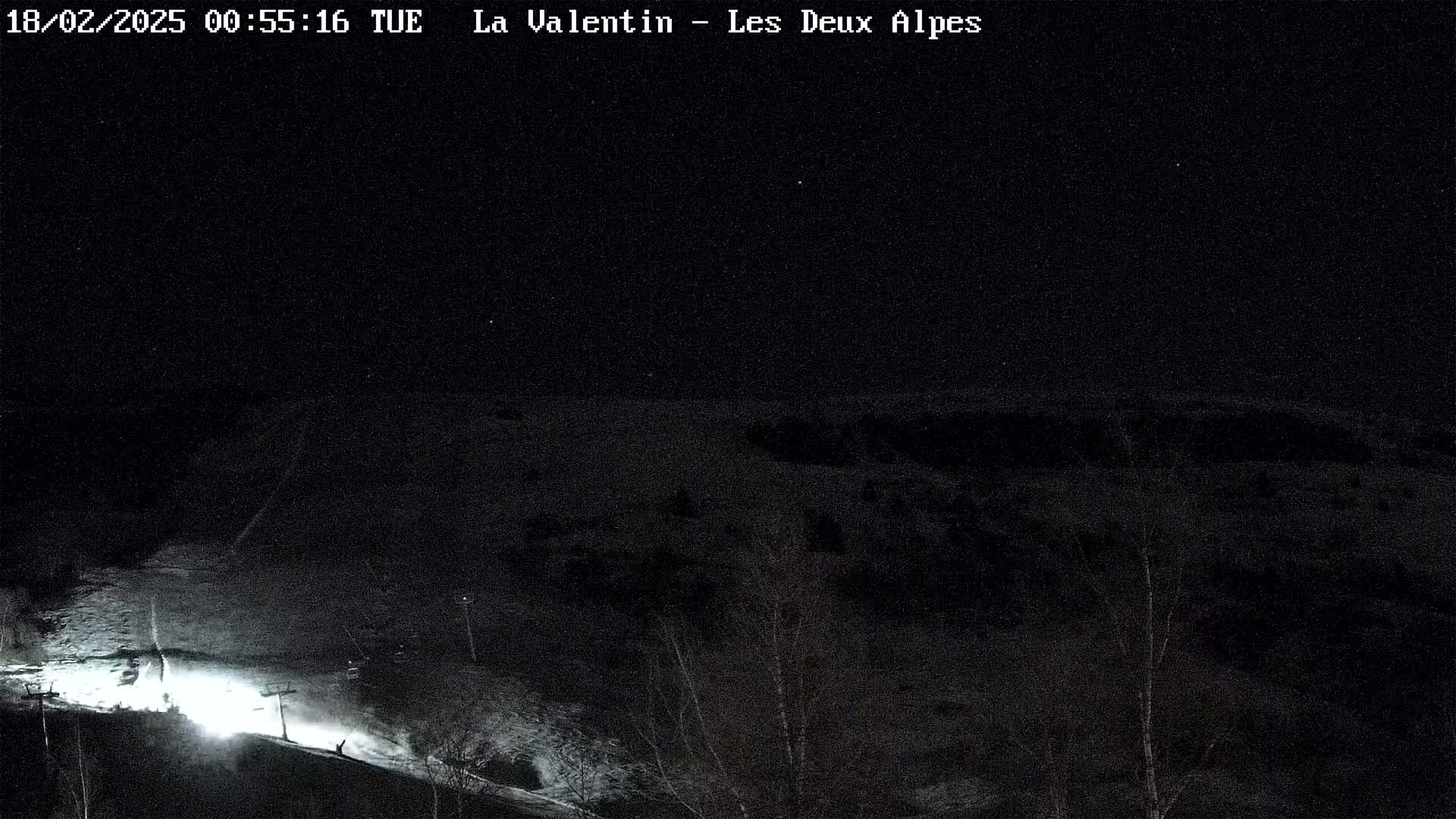A nighttime, snow-covered ski slope is illuminated by artificial light, with sparsely scattered trees visible in the background under a clear, dark sky.