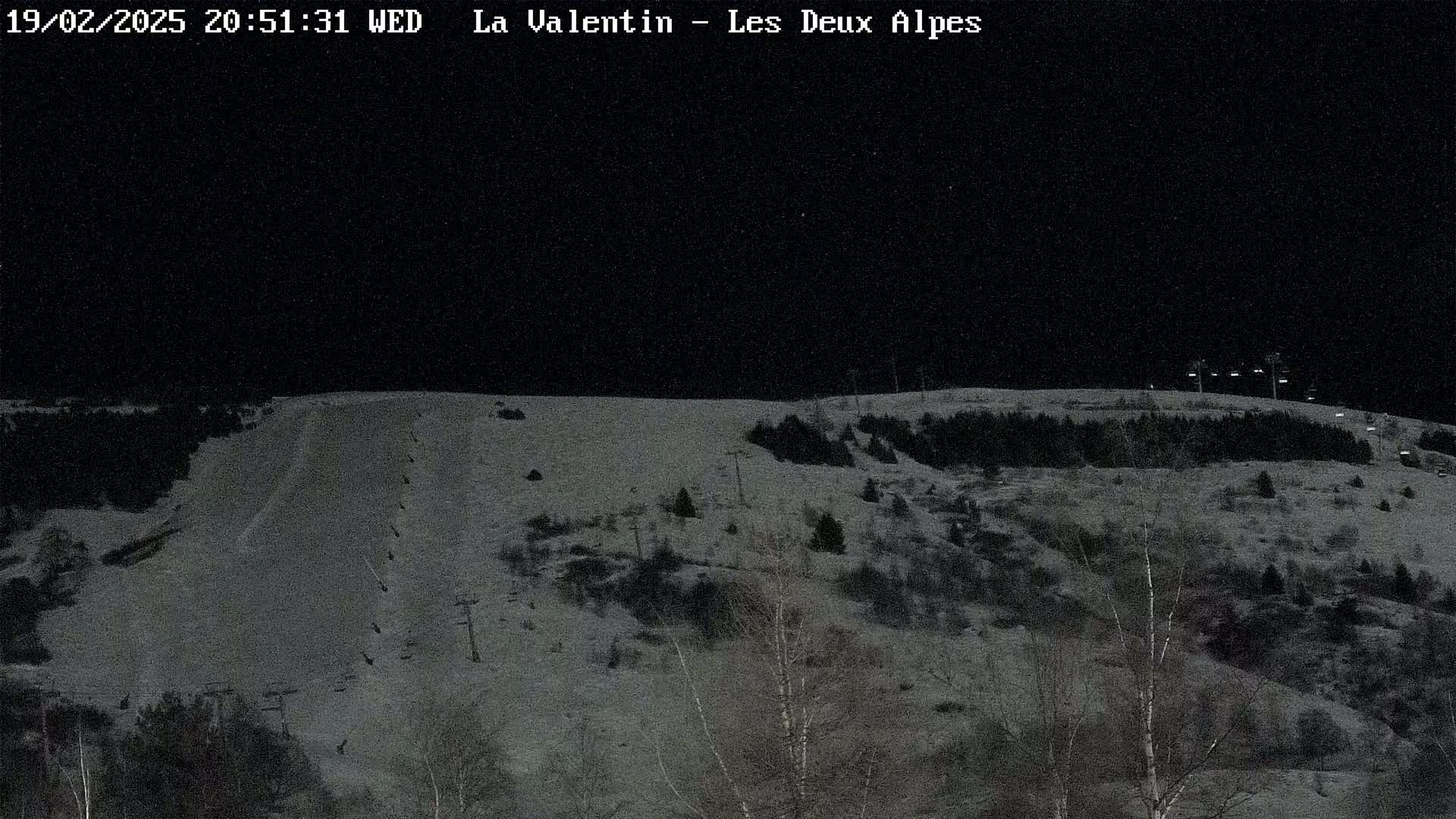 A snow-covered ski slope at night, illuminated by artificial lights, with sparsely wooded hillsides in the background under a dark sky.