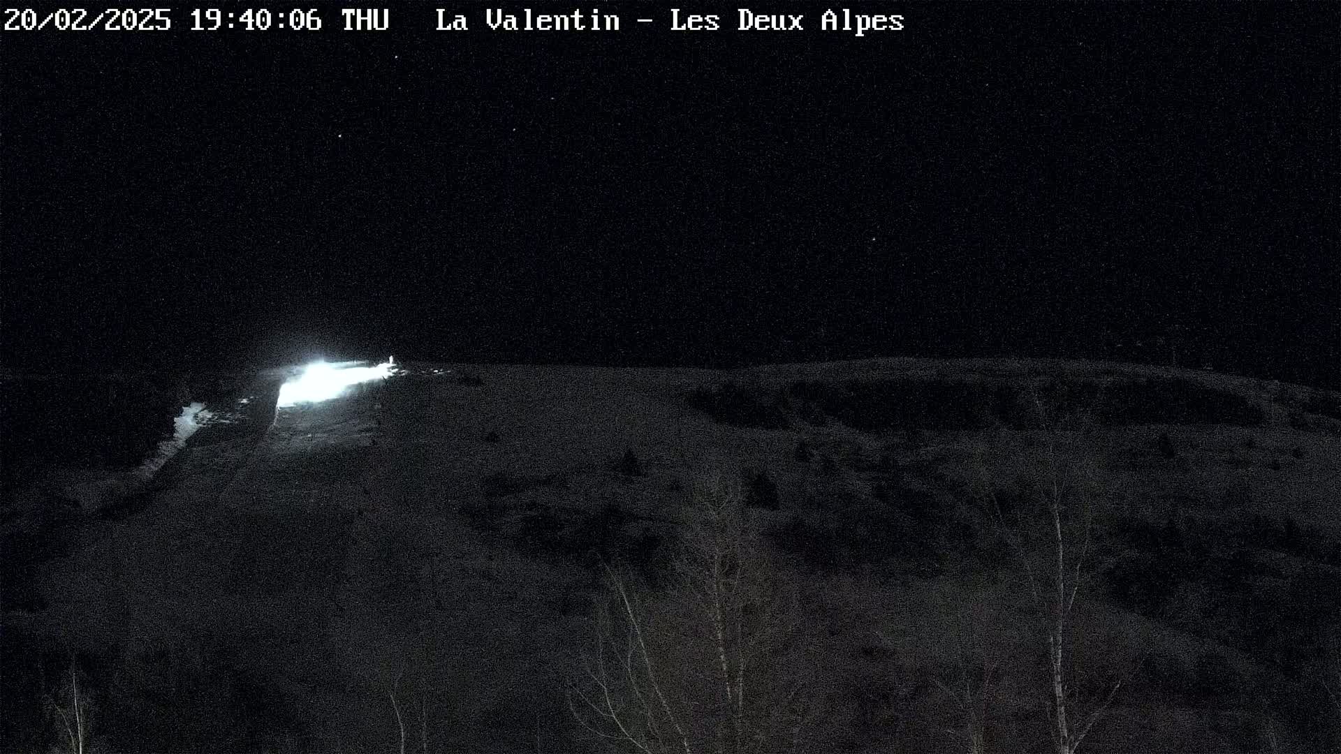A nighttime view of a snow-covered hillside illuminated by a bright light source in the distance, under a clear, starry sky.
