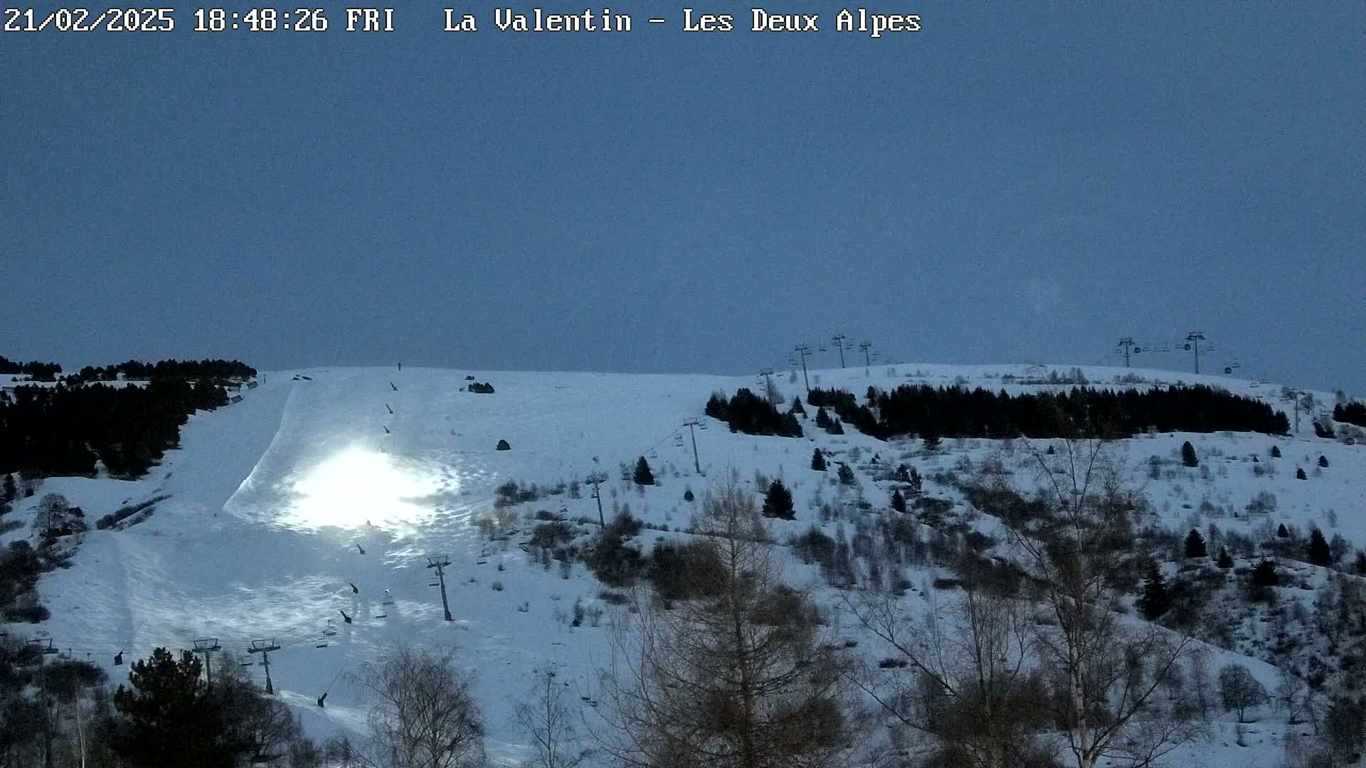 A snow-covered ski slope at dusk, illuminated by a bright light, with sparsely wooded hills and ski lifts visible.