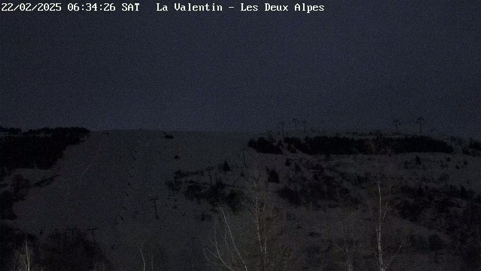 A dark, nighttime view of a snow-covered slope with sparse trees and ski lifts in the background under a dusky sky.